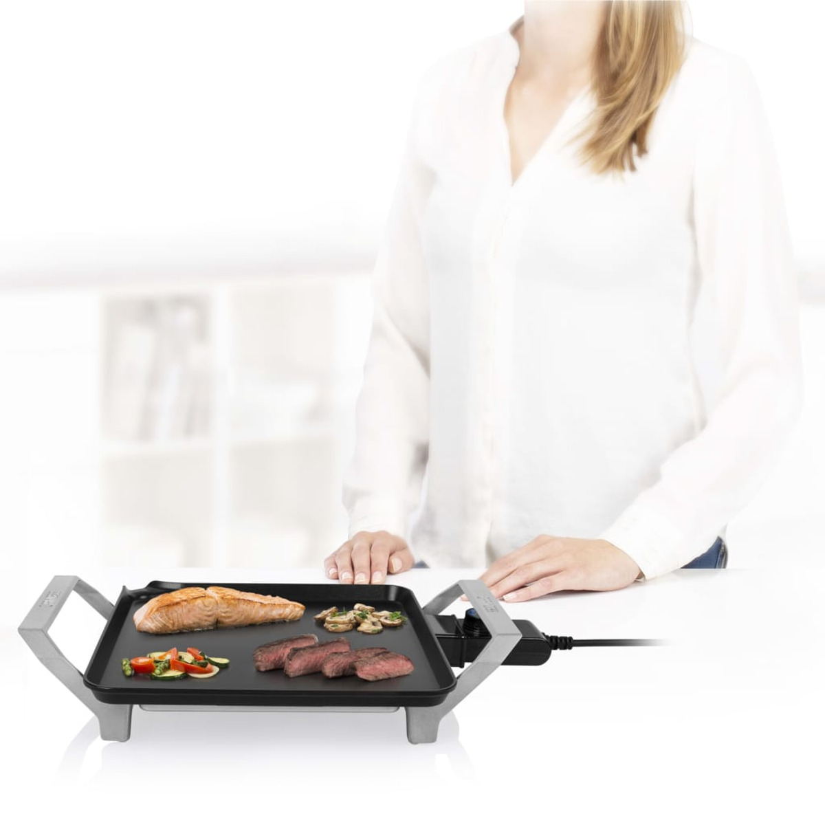 Mujer asando comida en una parrilla eléctrica negra sobre encimera blanca.