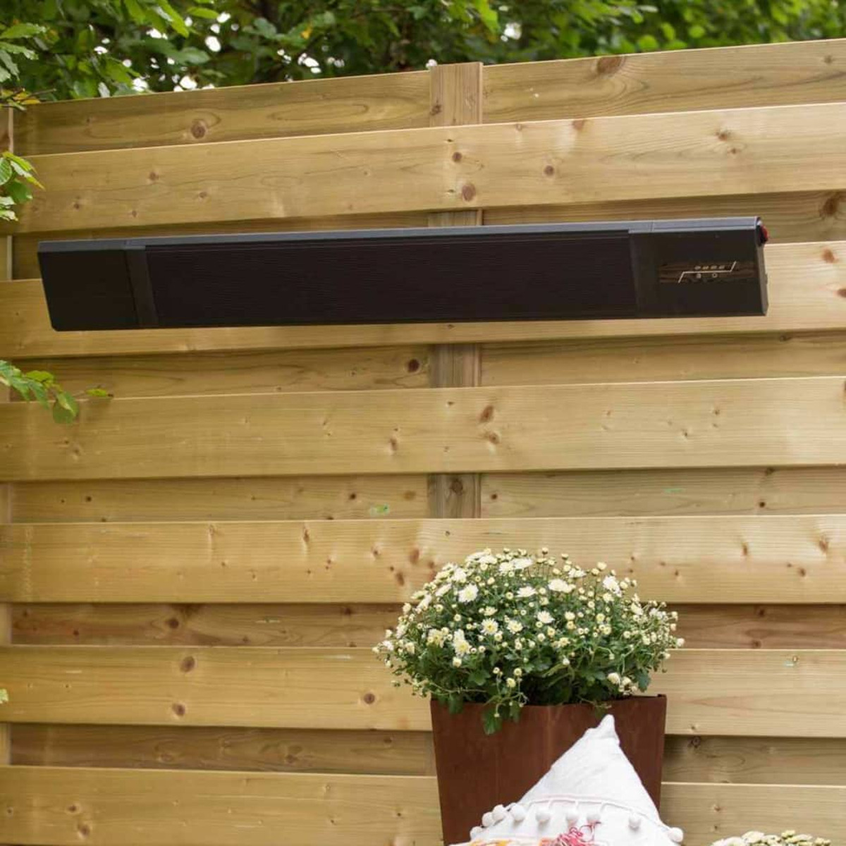 Black heater mounted on a wooden fence. White flowers in a brown pot below.