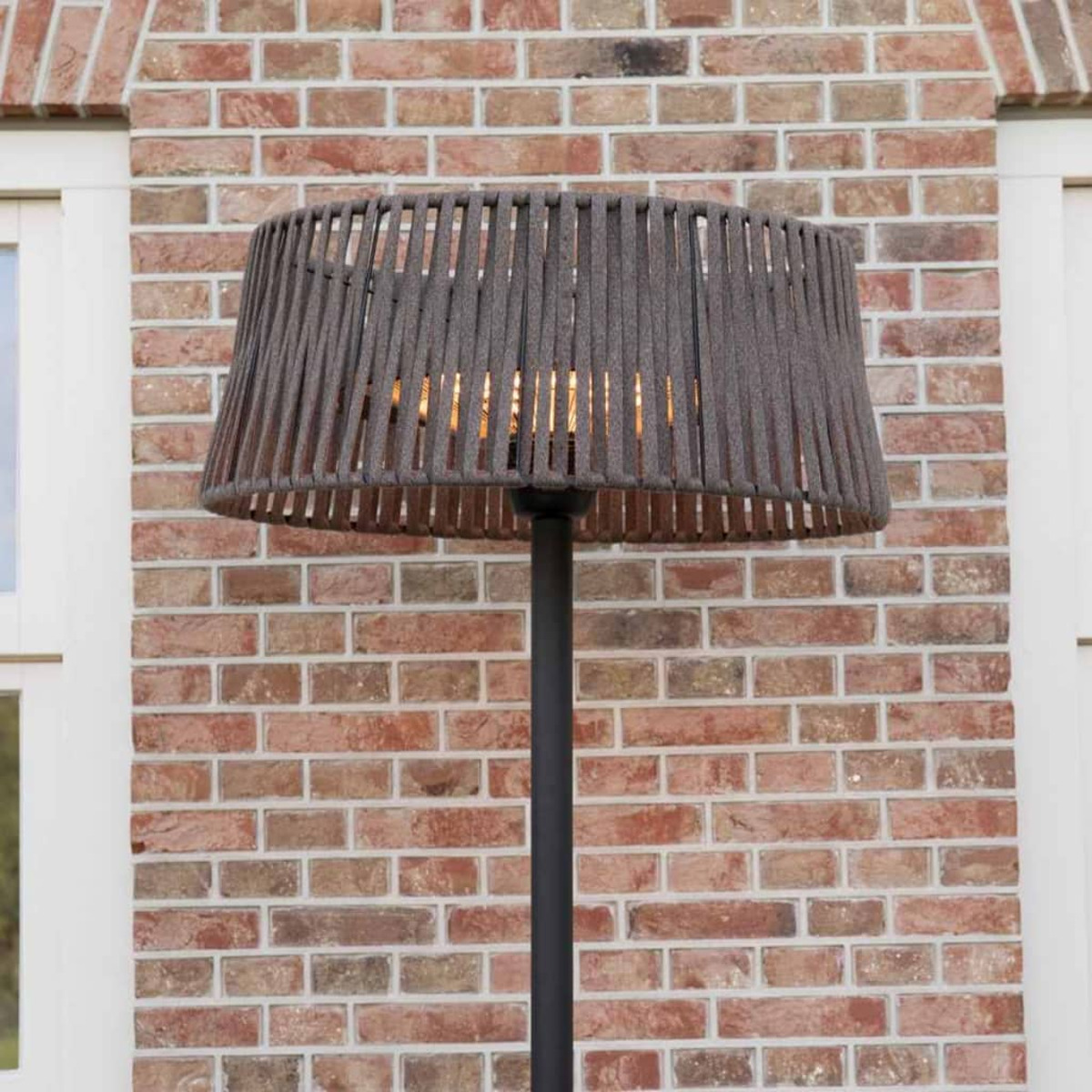 Een grijze buitenlamp met een geweven kap staat voor een bakstenen muur.