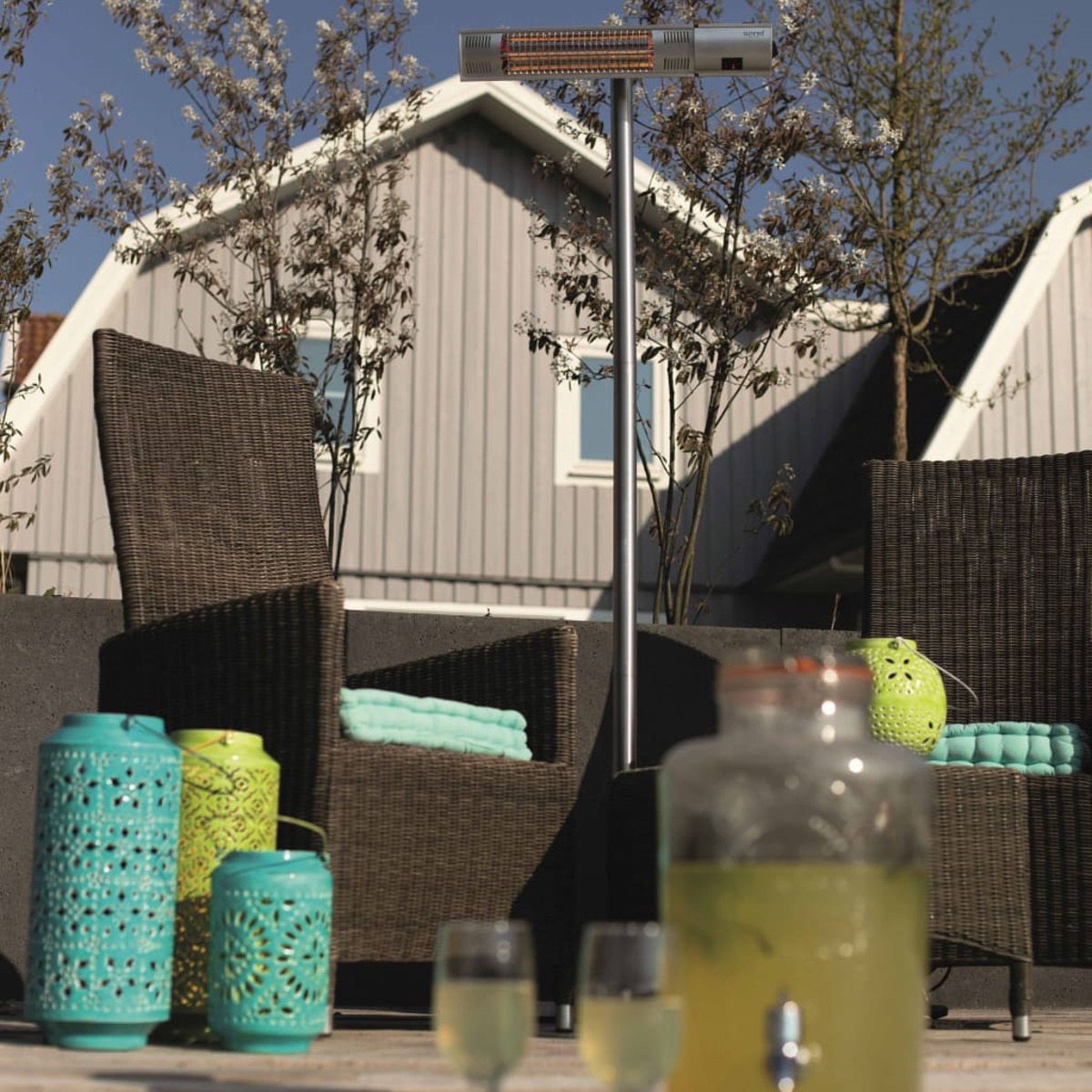Buitenscène met terrasmeubilair, lantaarns, drankjes en een staande heater. Huizen en bomen op de achtergrond.