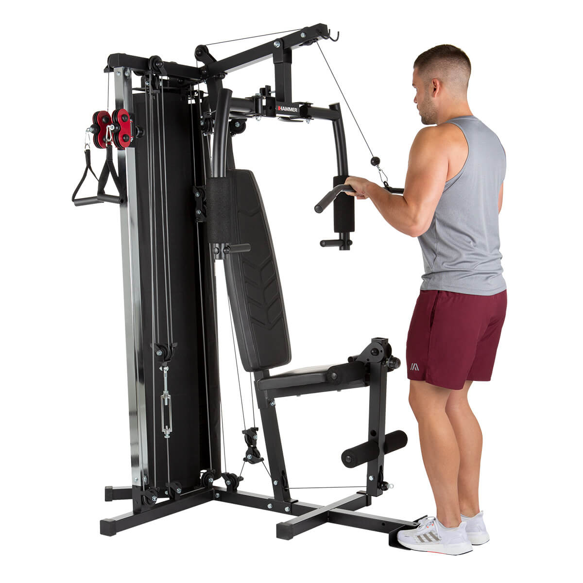 Mann benutzt ein Multigerät. Graues Tanktop und bordeauxrote Shorts. Schwarze und silberne Fitnessgeräte. Weißer Hintergrund.