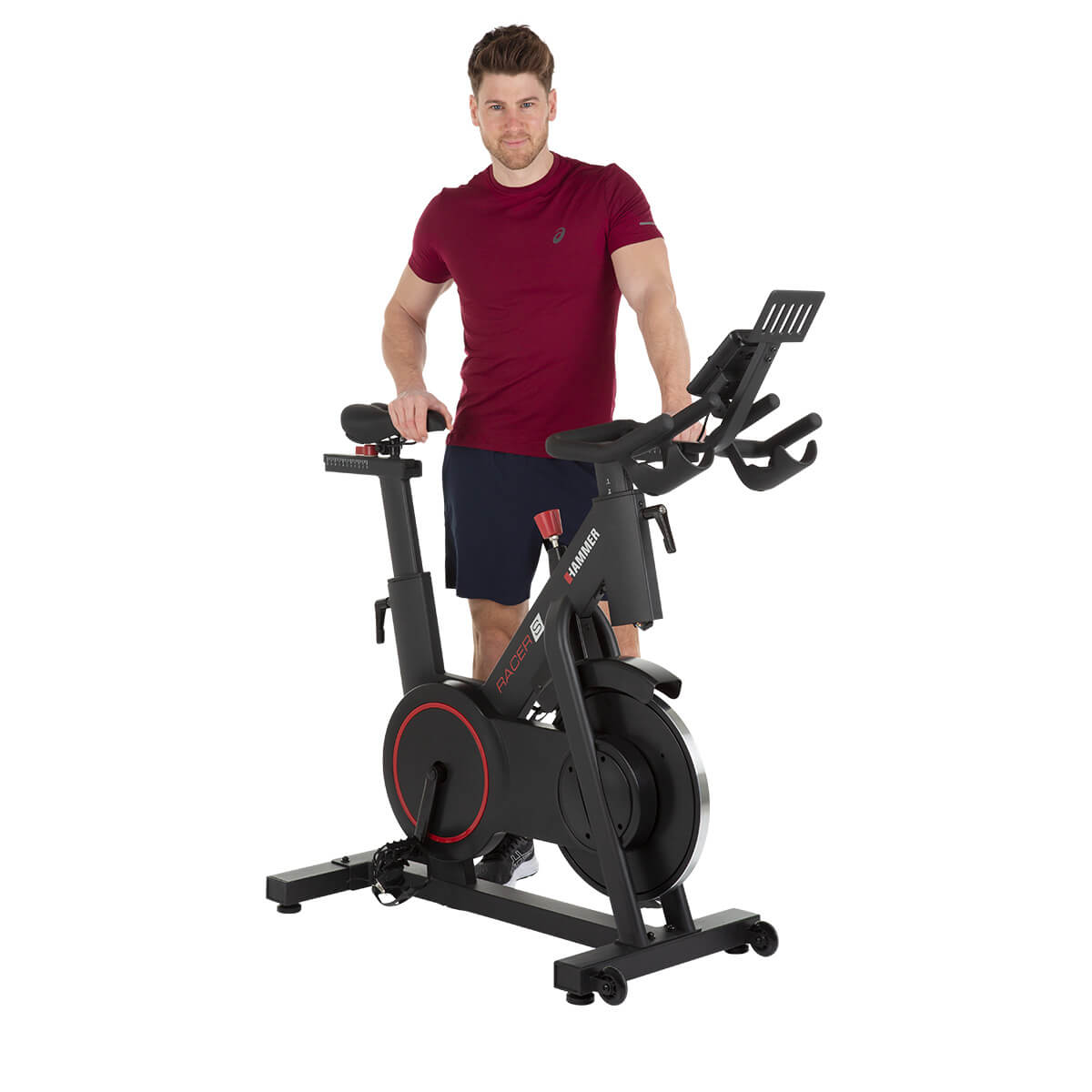 Mann steht neben einem schwarzen Heimtrainer, rotes Hemd und blaue Shorts, weißer Hintergrund.