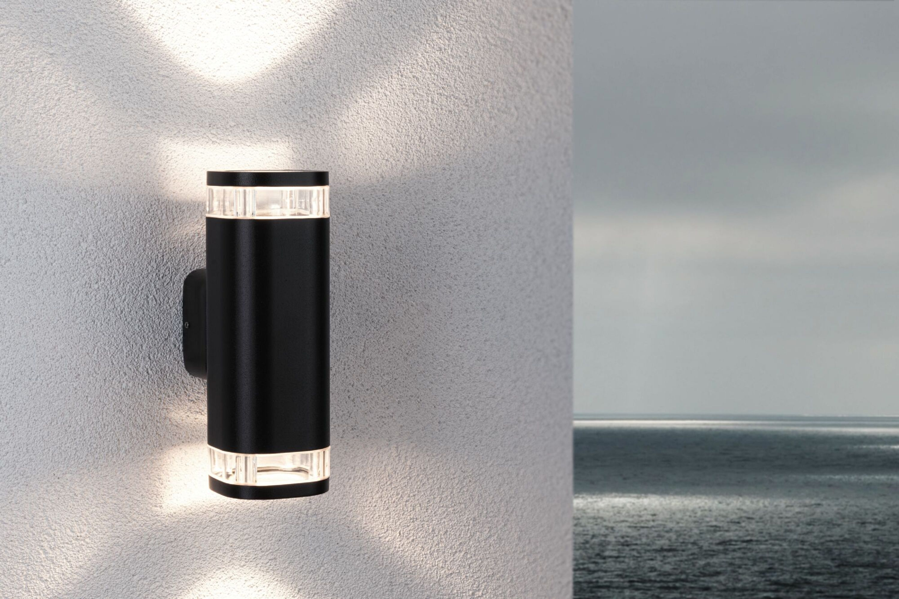 Schwarze Außenleuchte an einer weiß strukturierten Wand. Im Hintergrund ist das Meer zu sehen.