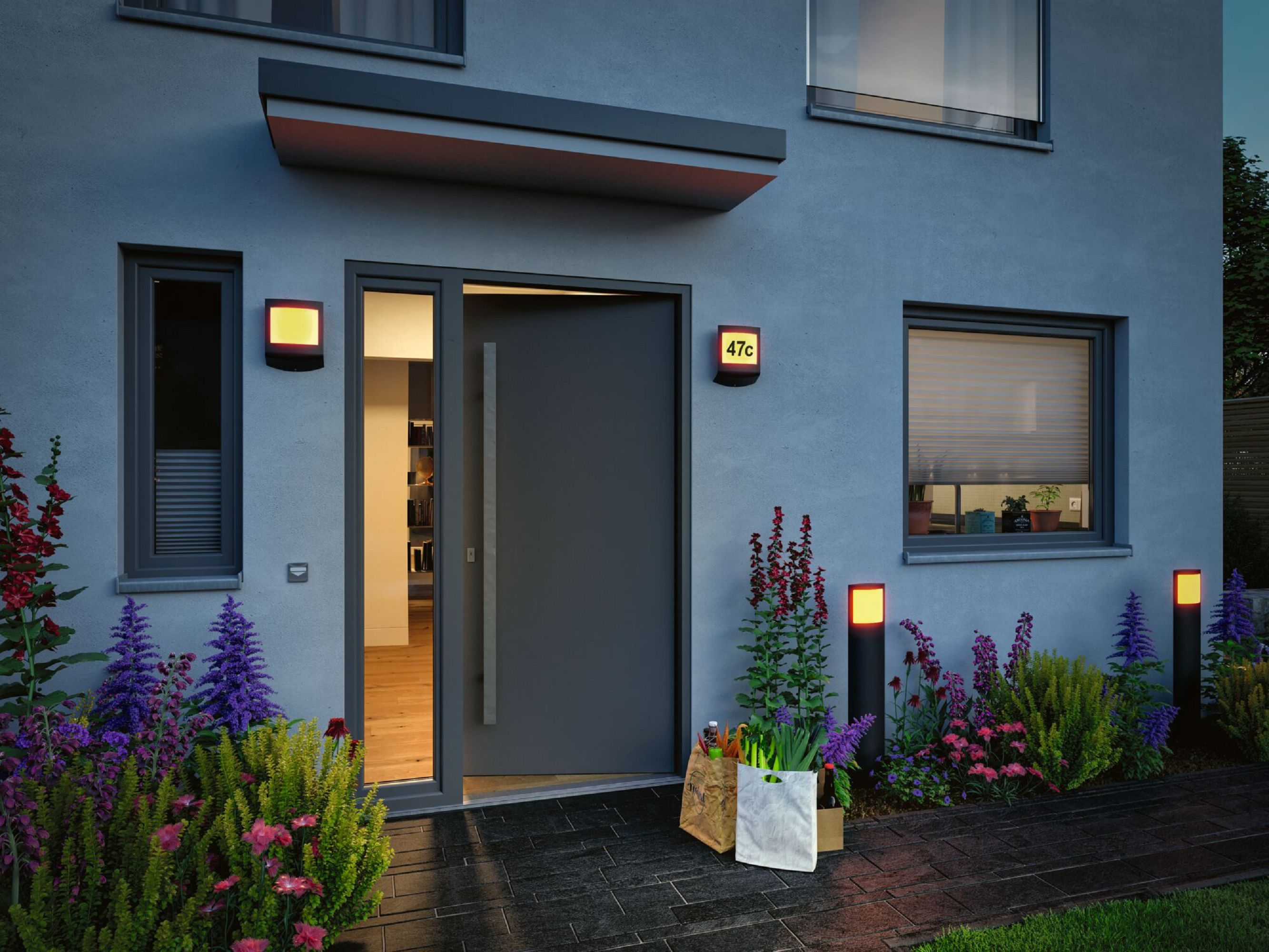 Zewnętrze domu w nocy z oświetlonym wejściem, światłami i rabatami kwiatowymi.