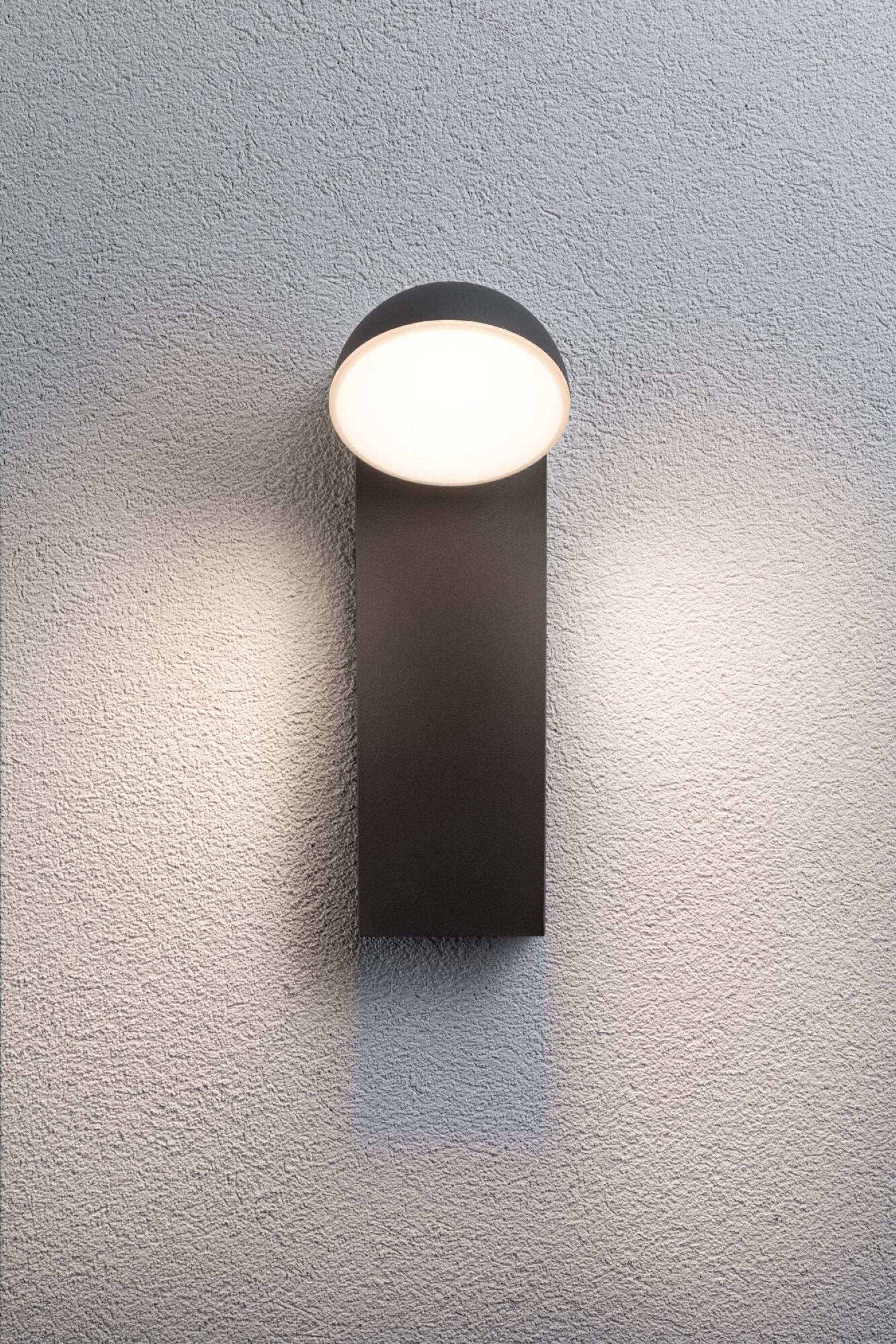 Eine schwarze, rechteckige Außenwandleuchte mit kreisförmiger Oberseite und weißer Lichtquelle, an der Wand.