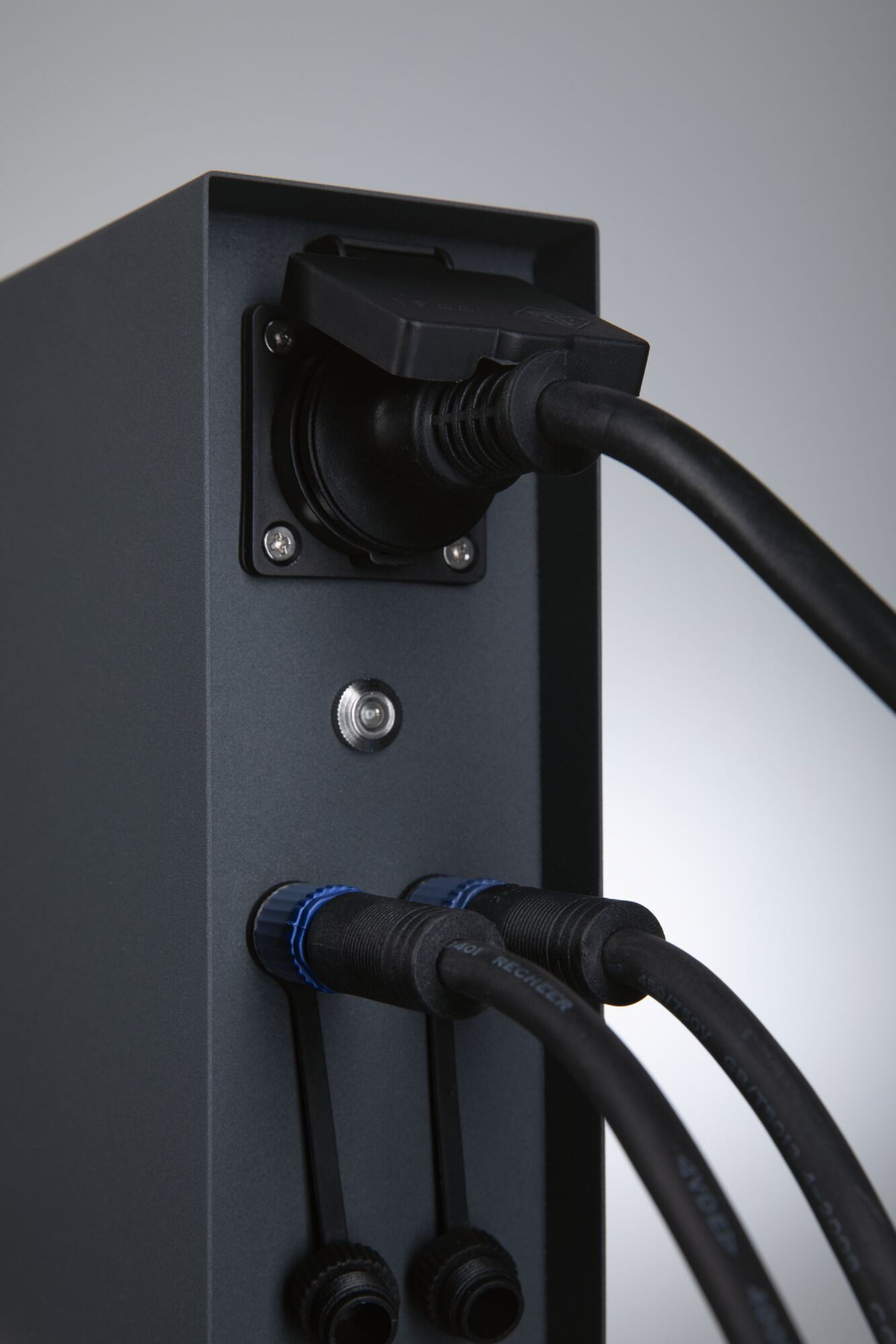 Schwarzer Metallkasten mit Stromkabeln und Steckern. Kabel sind eingesteckt. Grauer Hintergrund.