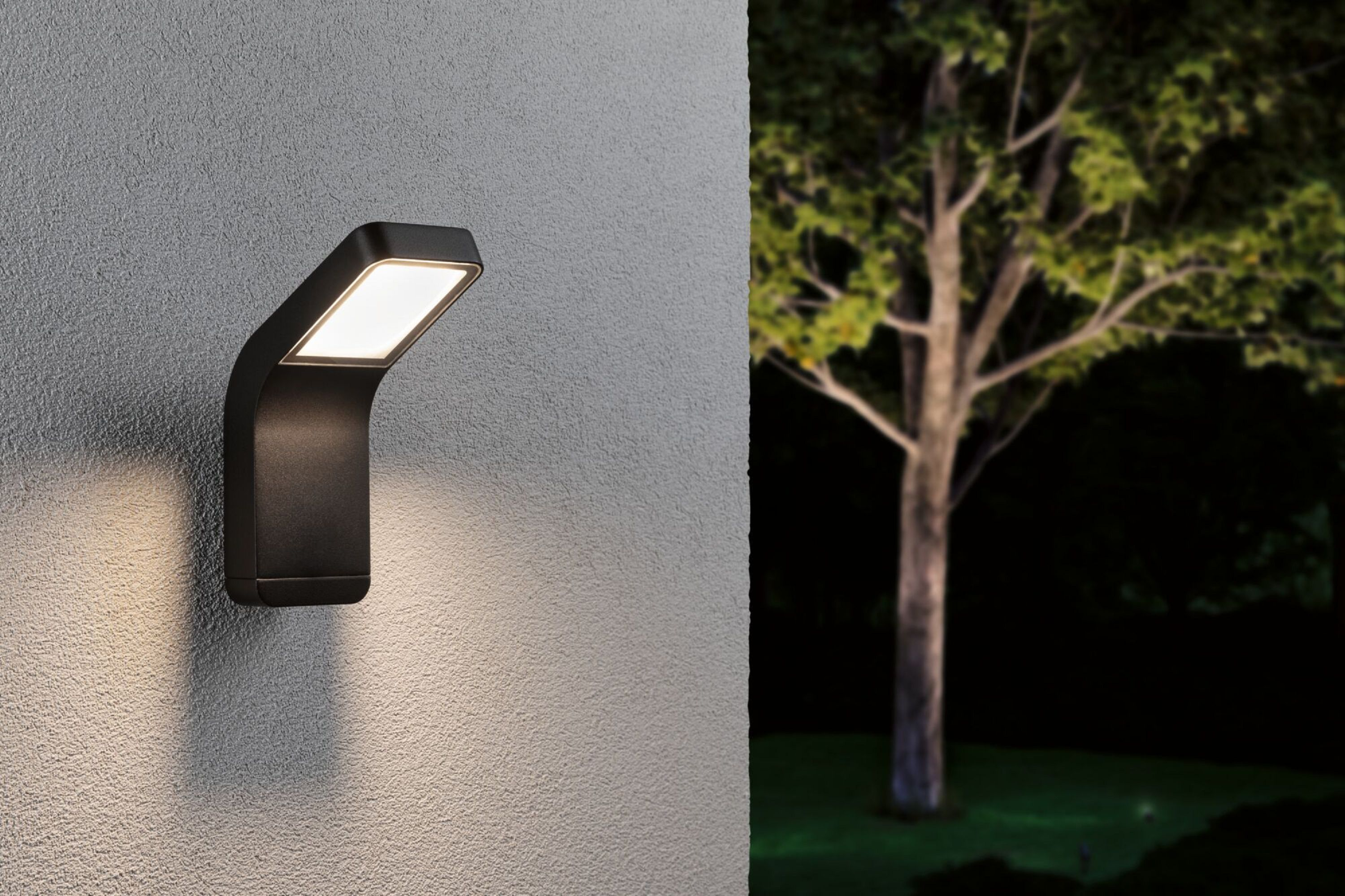 Eine schwarze, gebogene, rechteckige Außenleuchte ist an einer grauen Wand montiert. Ein Baum ist im Hintergrund.