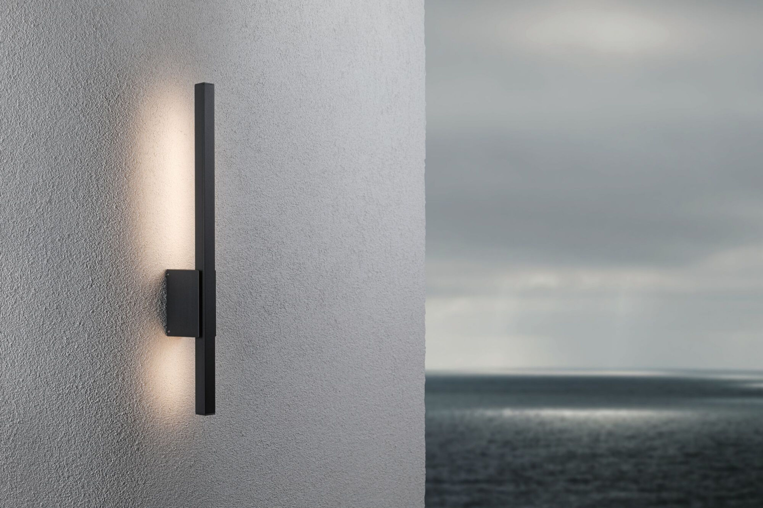Eine Außenwandleuchte an einer strukturierten Wand mit Blick auf das Meer im Hintergrund.