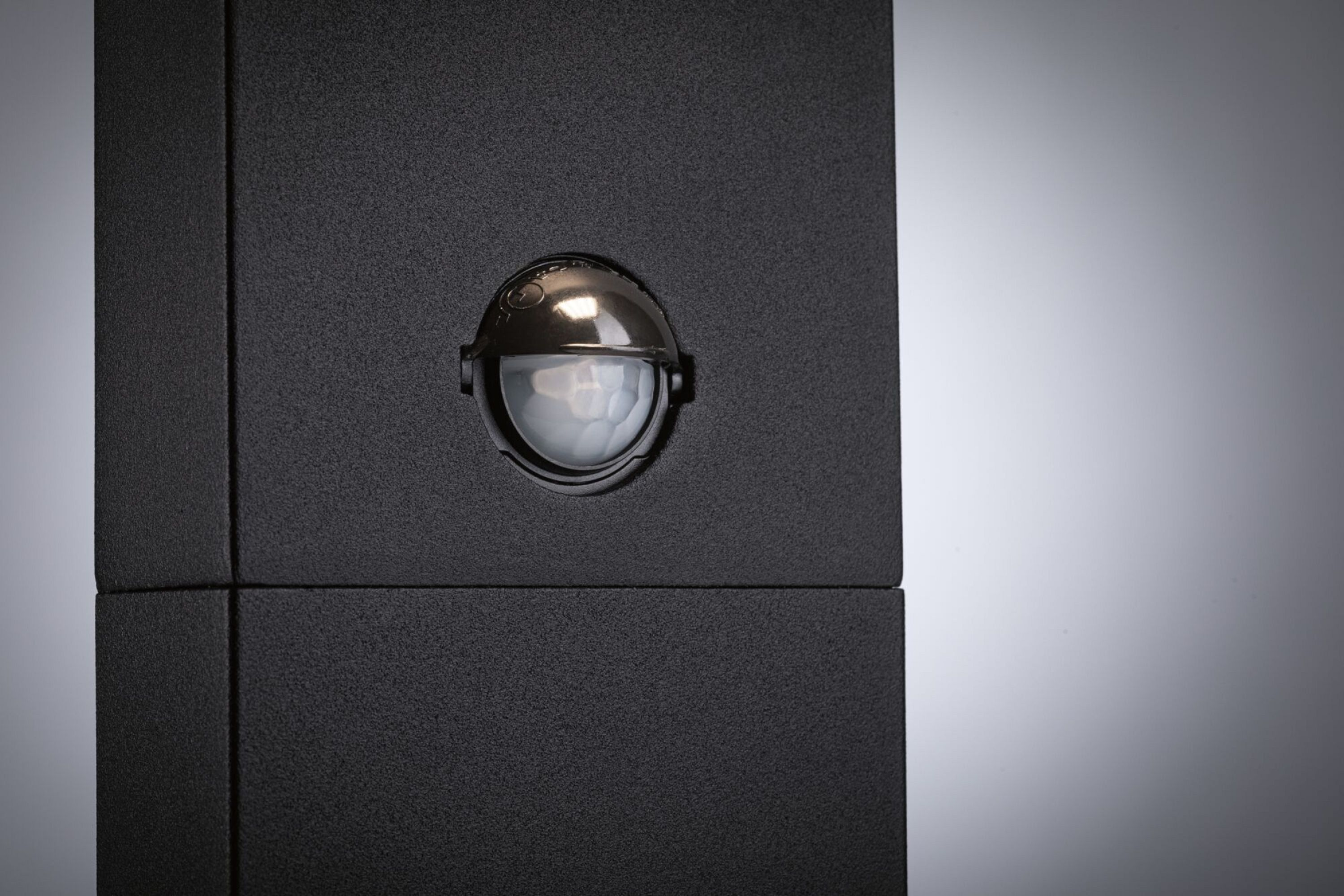 Schwarzer Sensor auf schwarzer Oberfläche. Der Sensor ist rund, mit transparenter Kuppel. Weißer Hintergrund.