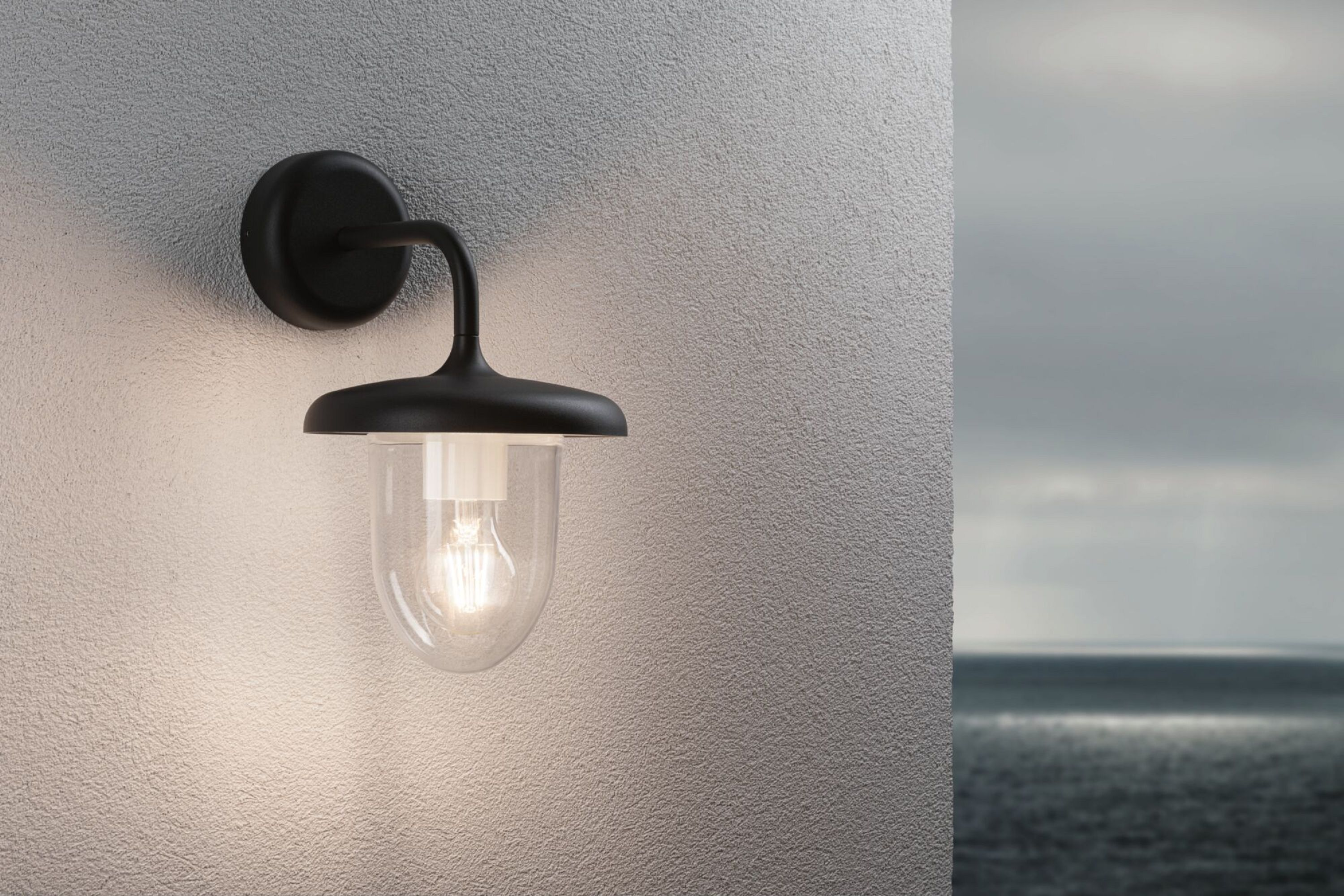 Schwarze Außenlampe an grauer Wand, Glühbirne leuchtet. Meer im Hintergrund.