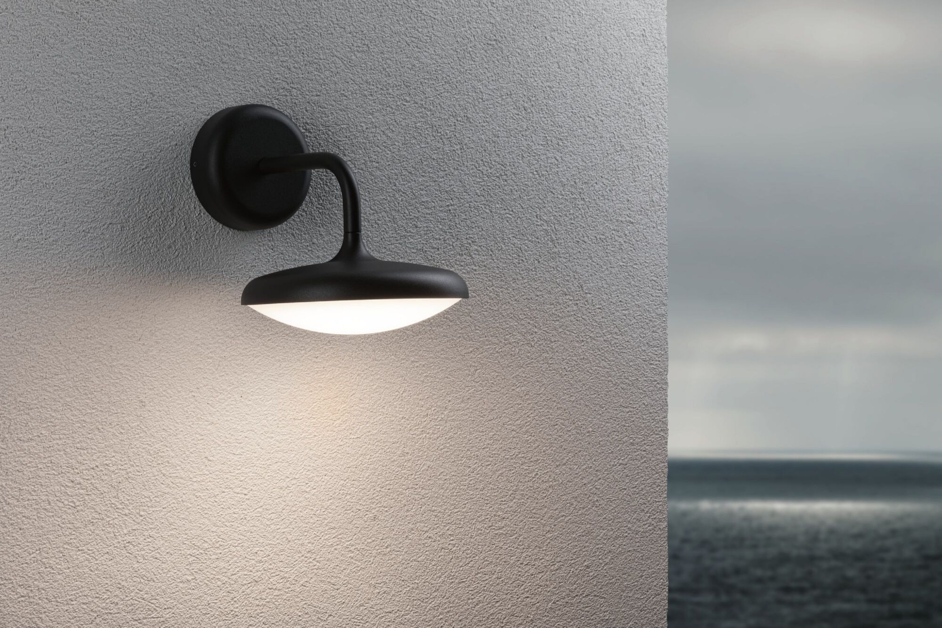 Schwarze Wandleuchte an einer strukturierten grauen Wand. Die Lampe hat einen gebogenen Arm und rundes Licht. Verschwommener Hintergrund.