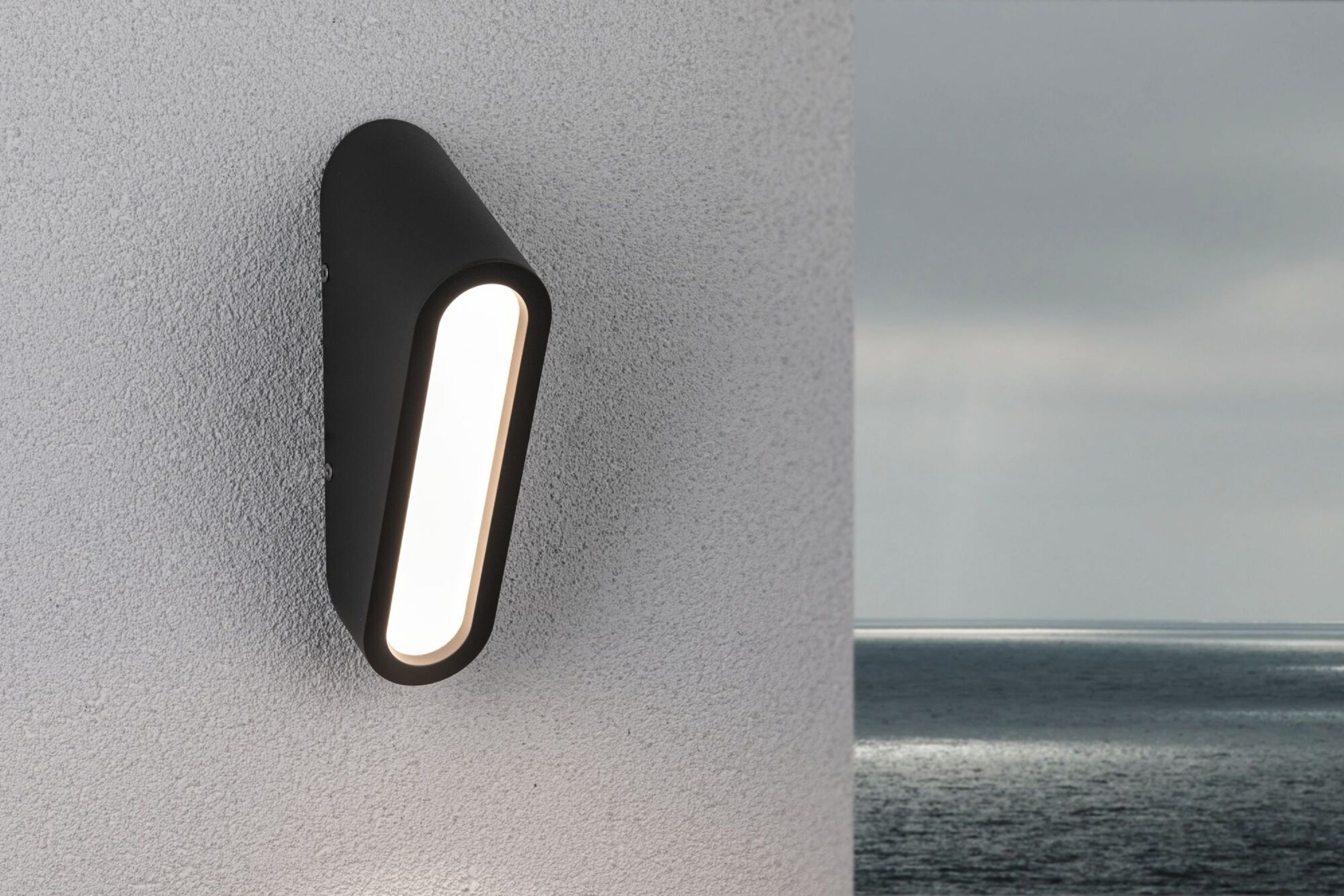 Schwarze Außenwandleuchte. Licht ist an. Der Hintergrund ist ein bewölkter Himmel und Wasser.
