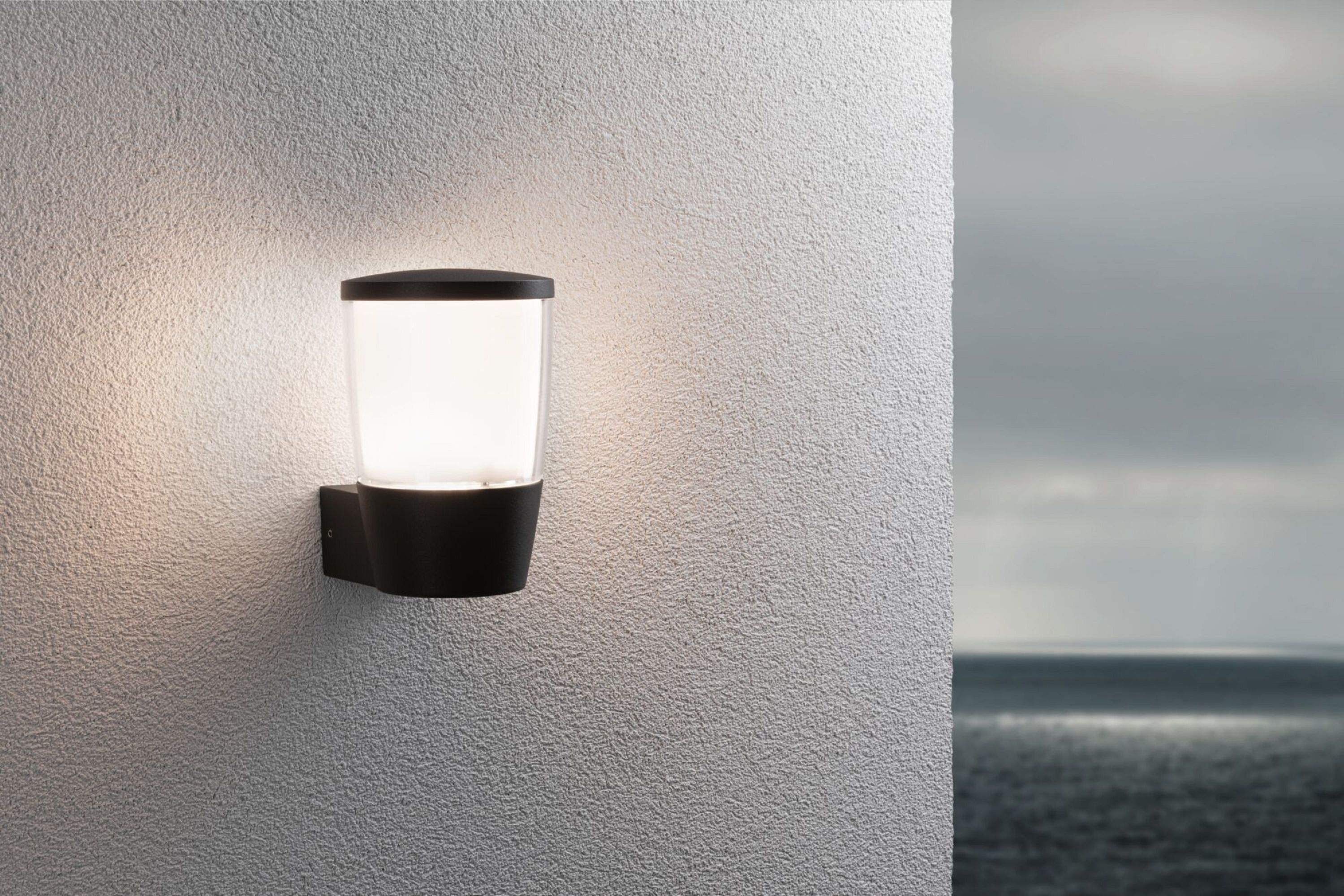 Schwarze Außenwandleuchte mit warmem Licht an strukturierter weißer Wand, unscharfer Hintergrund.