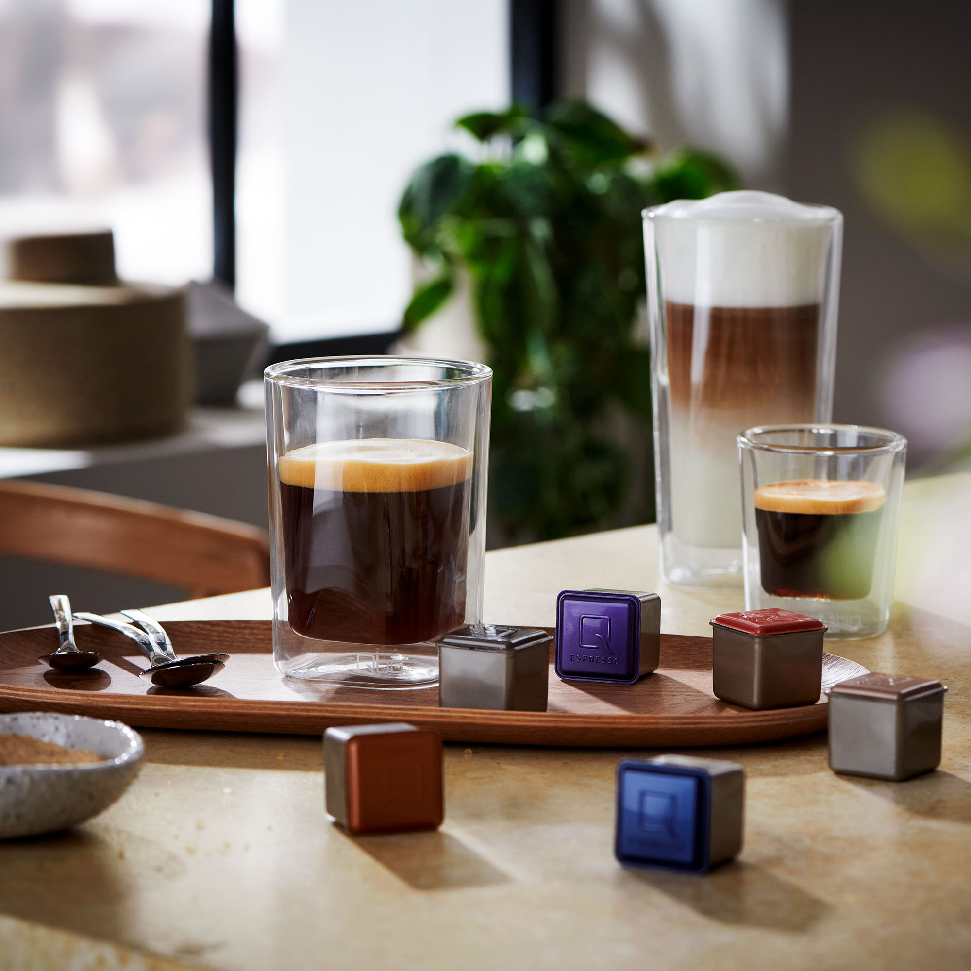 Kaffee- und Cappuccino-Getränke, Kaffeekapseln und ein Holztablett auf einem Tisch mit Pflanzen im Hintergrund.