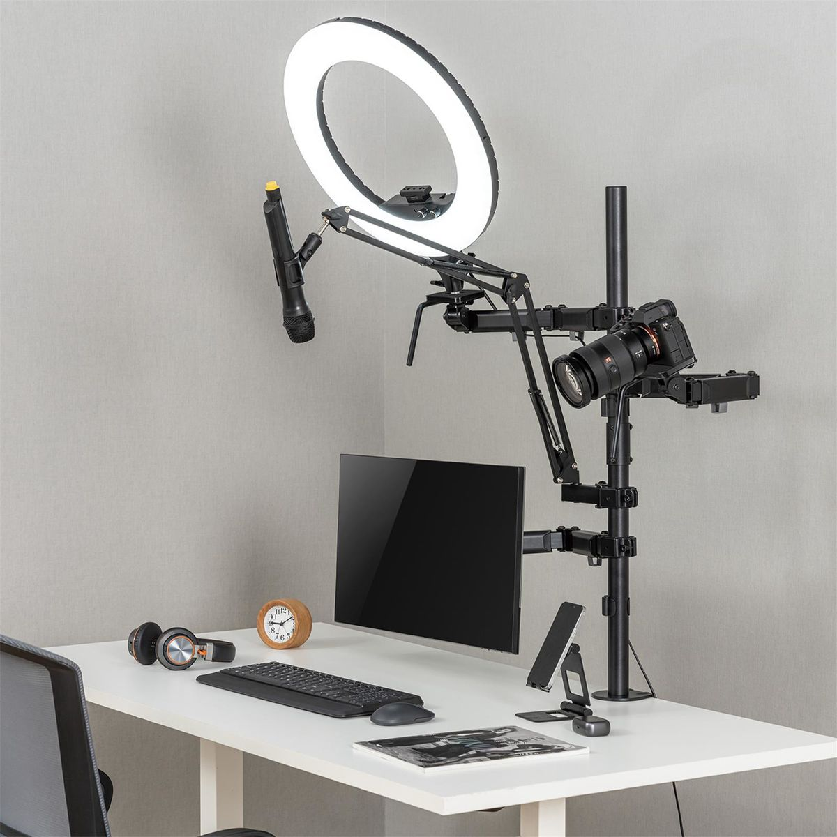 Bureau-opstelling met ringlicht, microfoon, camera, monitor, toetsenbord, muis, koptelefoon en klok.