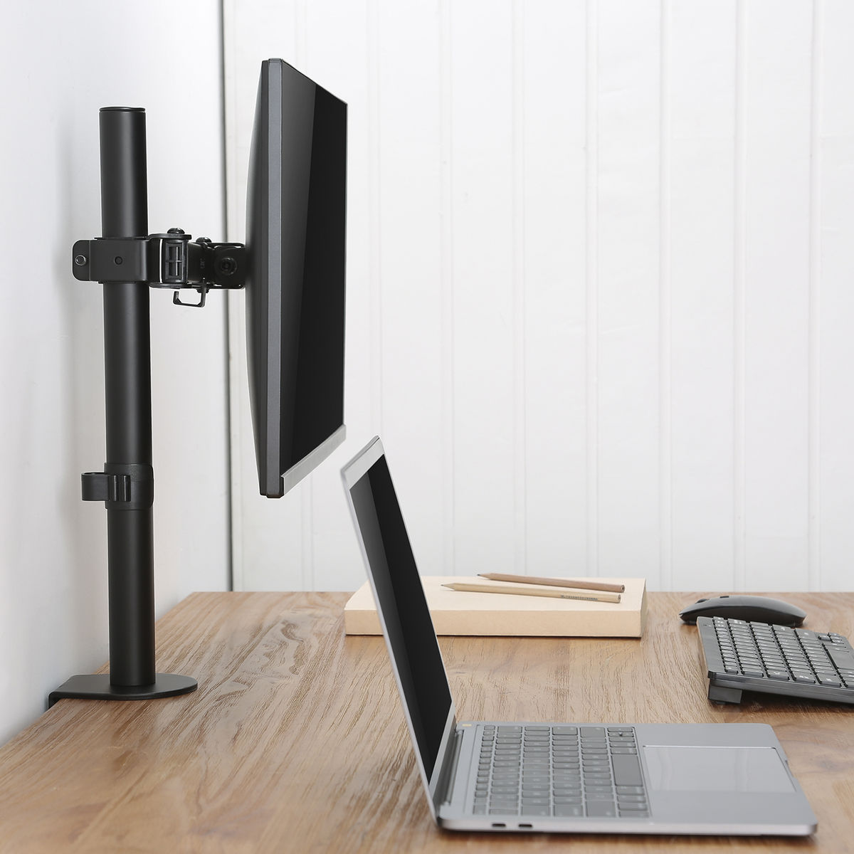Een computermonitor gemonteerd op een zwarte arm, met een laptop en accessoires op een bureau.