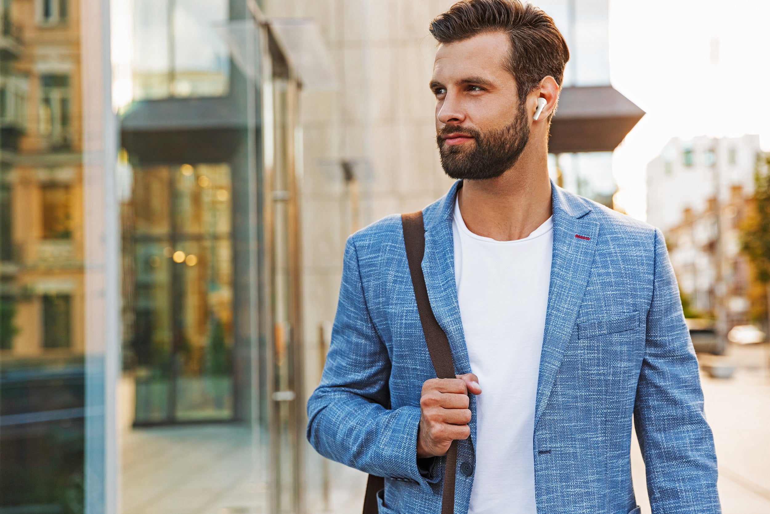 Uomo barbuto con blazer blu e auricolari. Porta una borsa ed è all'aperto.