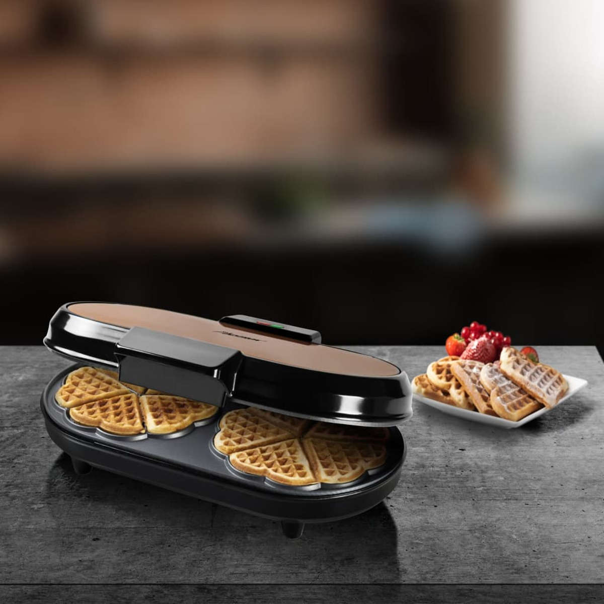 Macchina per waffle con waffle e un piatto di fragole e altri waffle, sfondo scuro.