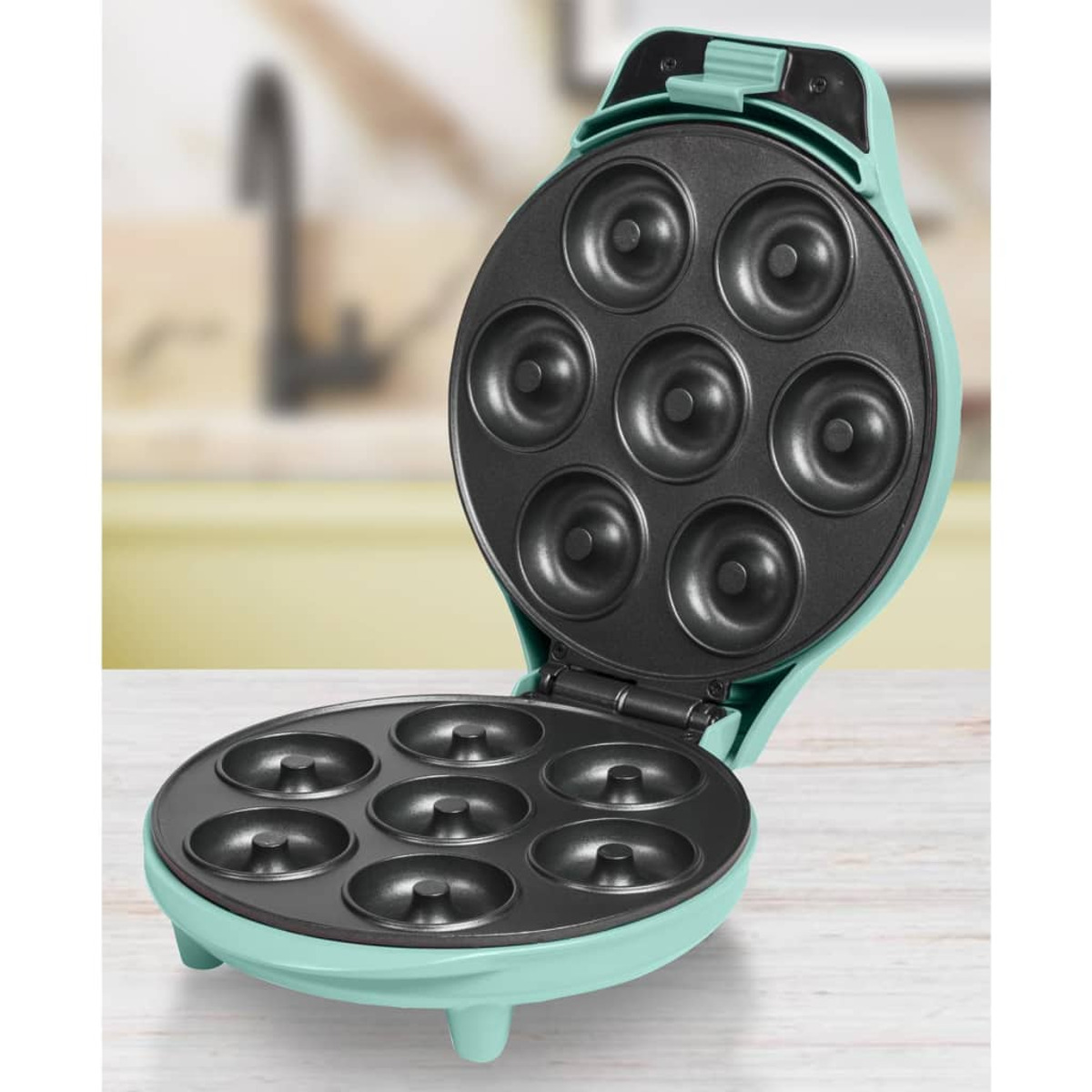 Máquina de hacer donas turquesa abierta, revelando siete moldes. Fondo de cocina borroso.
