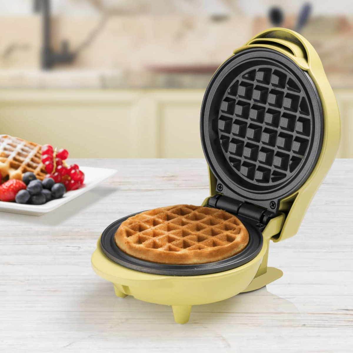 Una máquina para hacer gofres amarilla, abierta, con un gofre cocido dentro. Un plato con gofres, bayas y fruta al lado.