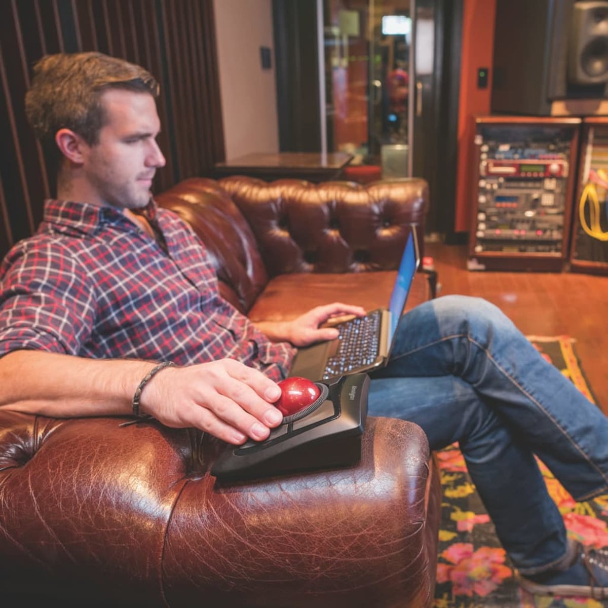 A man sitting on a leather couch using a laptop with a trackball mouse.