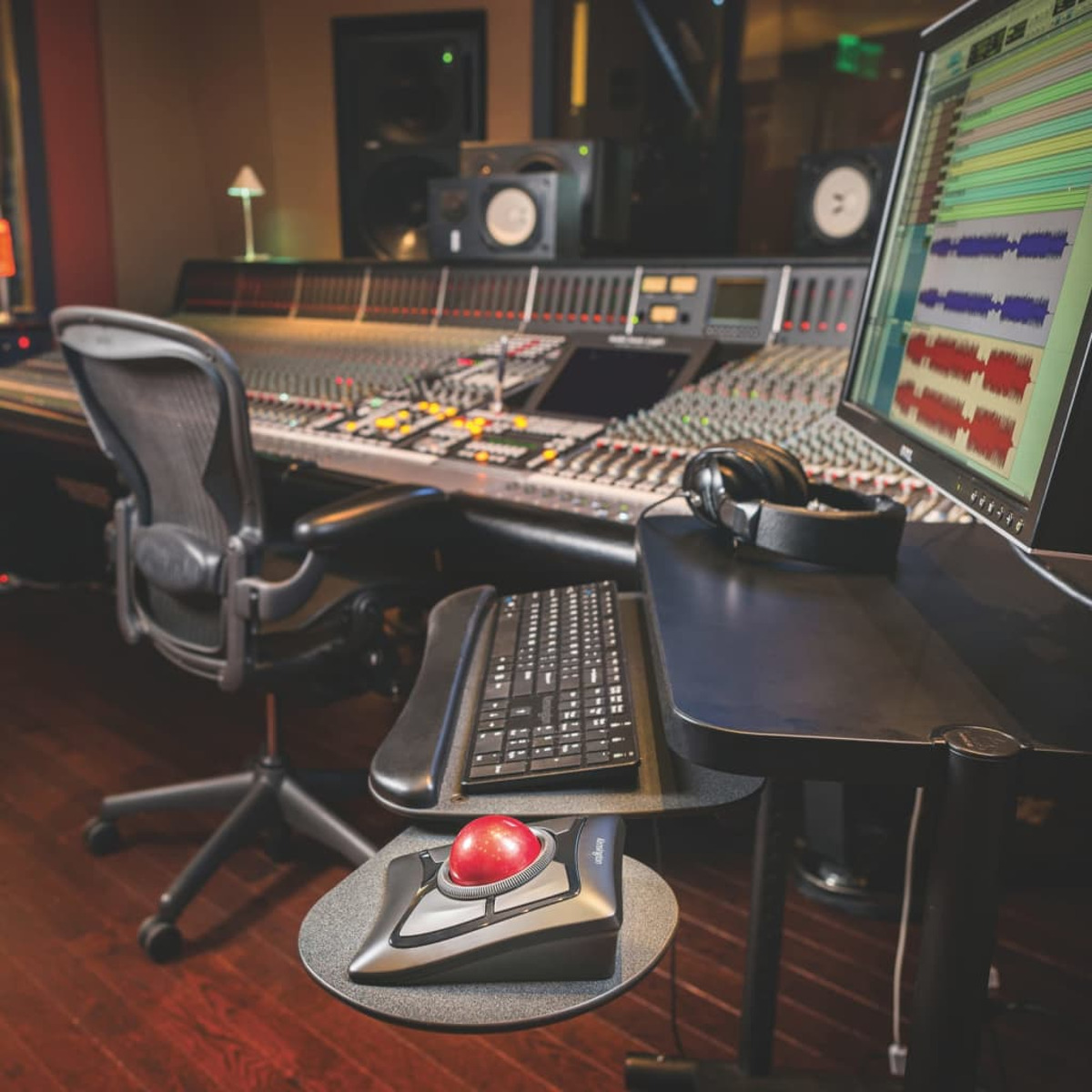 A sound studio with a mixing console, computer monitor, keyboard, and a red trackball mouse.