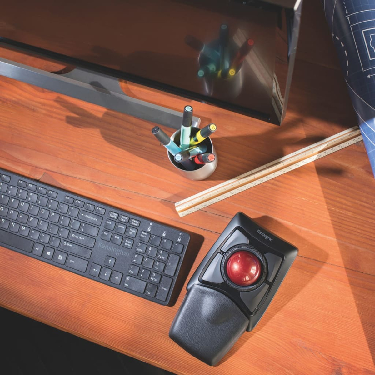 Desk setup with a keyboard, trackball mouse, pens, and monitor. Wooden desk.