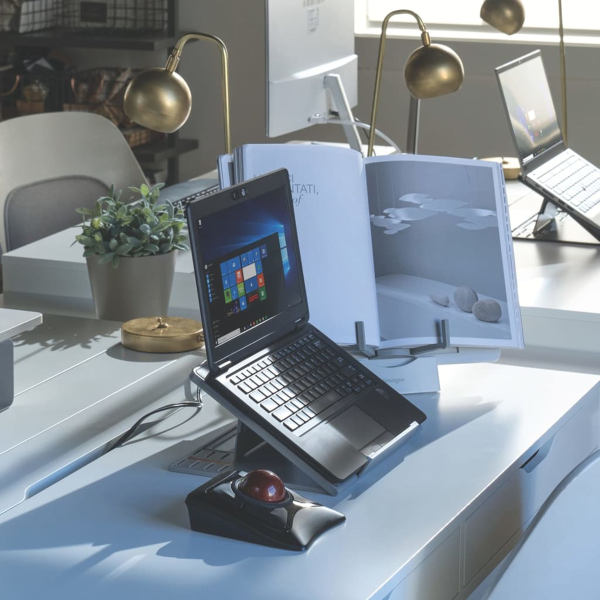 Office desk with a laptop, trackball mouse, and open book. Two gold lamps are visible.