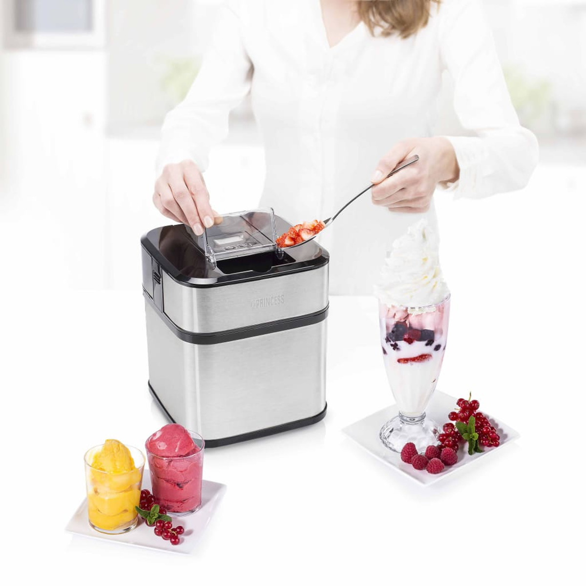 Kobieta dodaje truskawki do maszyny do lodów, z gotowymi pucharkami i lodami z boku.