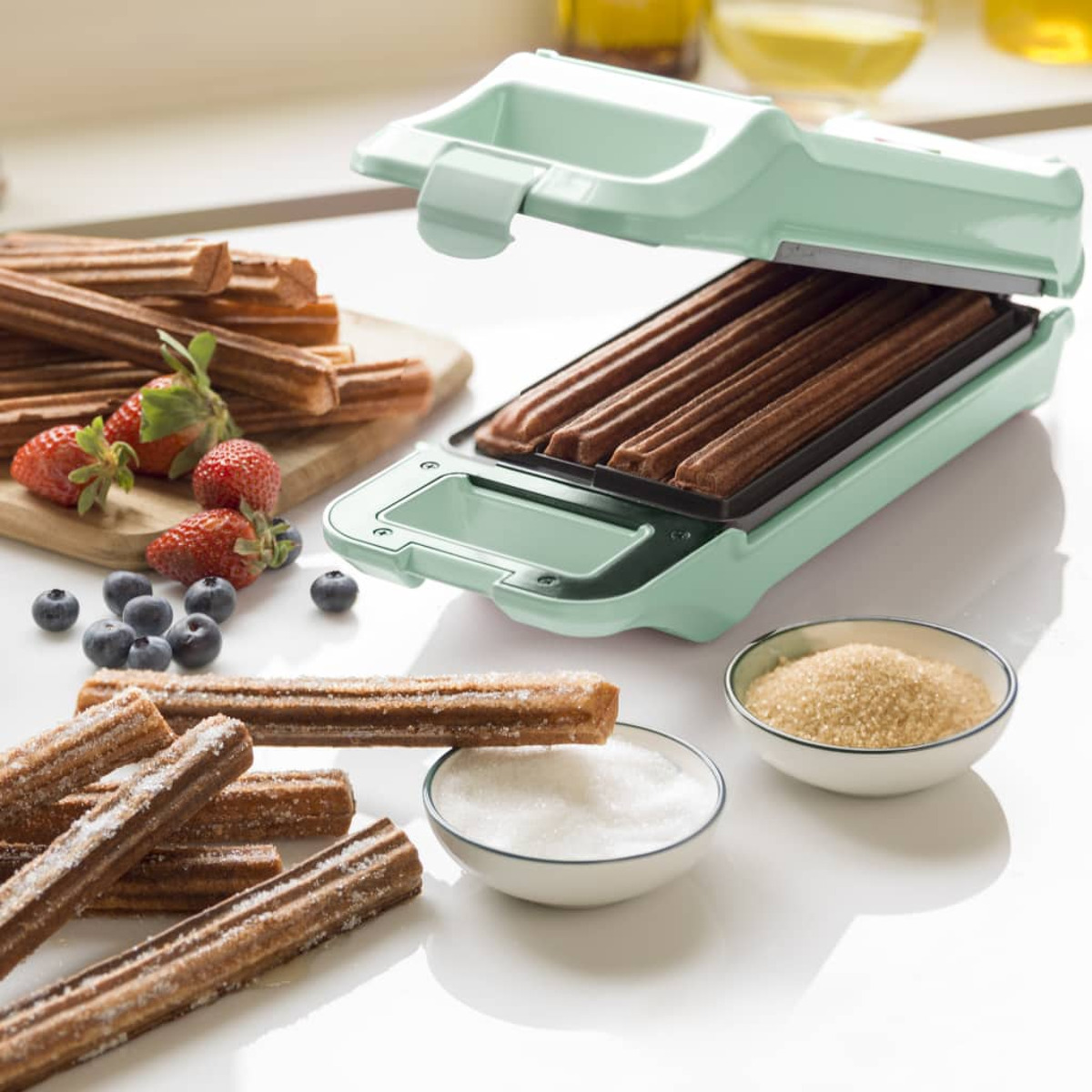 Macchina per churros con churros freschi, zucchero e frutti di bosco su superficie bianca.