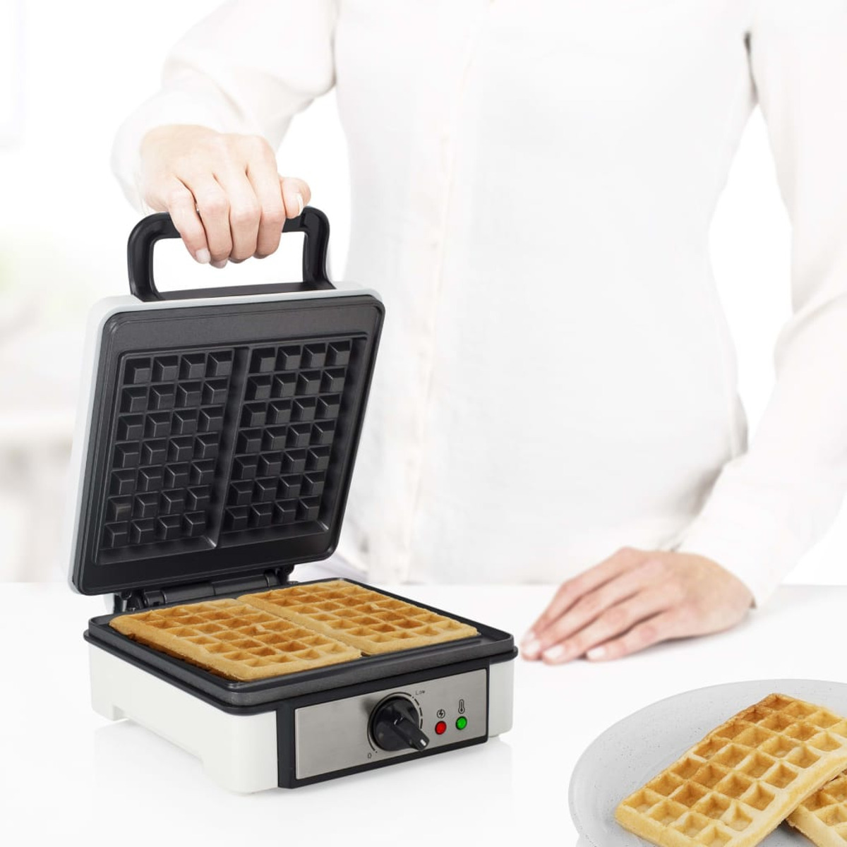 Kobieta obsługuje gofrownicę. Urządzenie jest otwarte, pokazując gofry w środku. Gofry na talerzu.