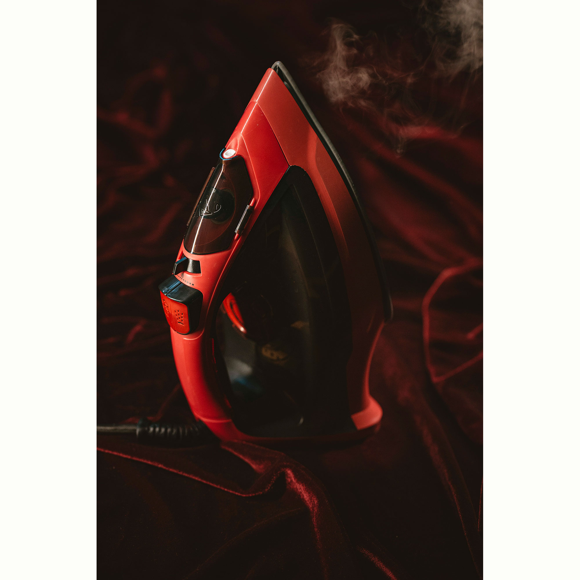 Czerwono-czarne żelazko z parą unoszącą się na tle ciemnoczerwonej tkaniny.