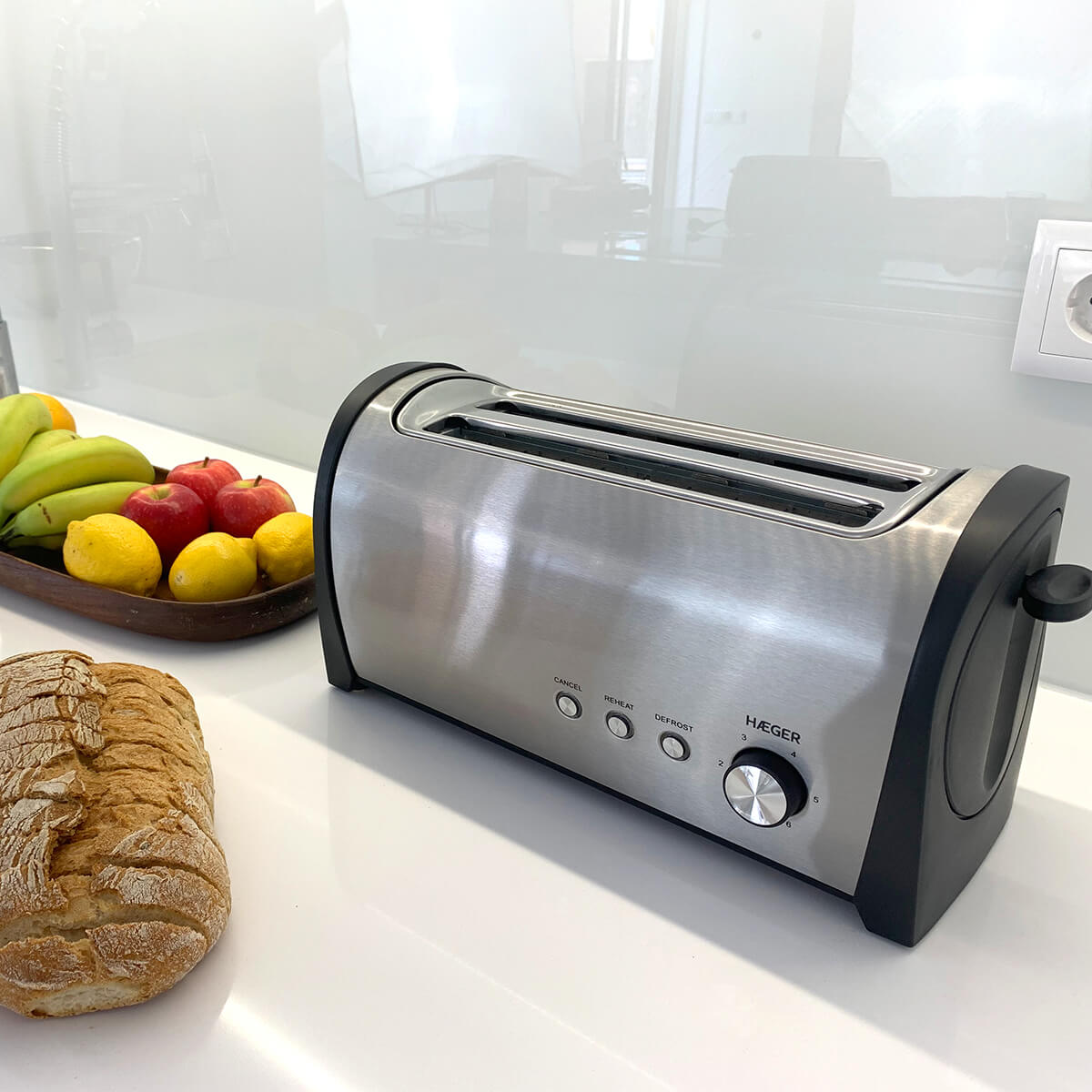 Srebrny toster z czarnymi akcentami, bochenek chleba i miska owoców na białym blacie.