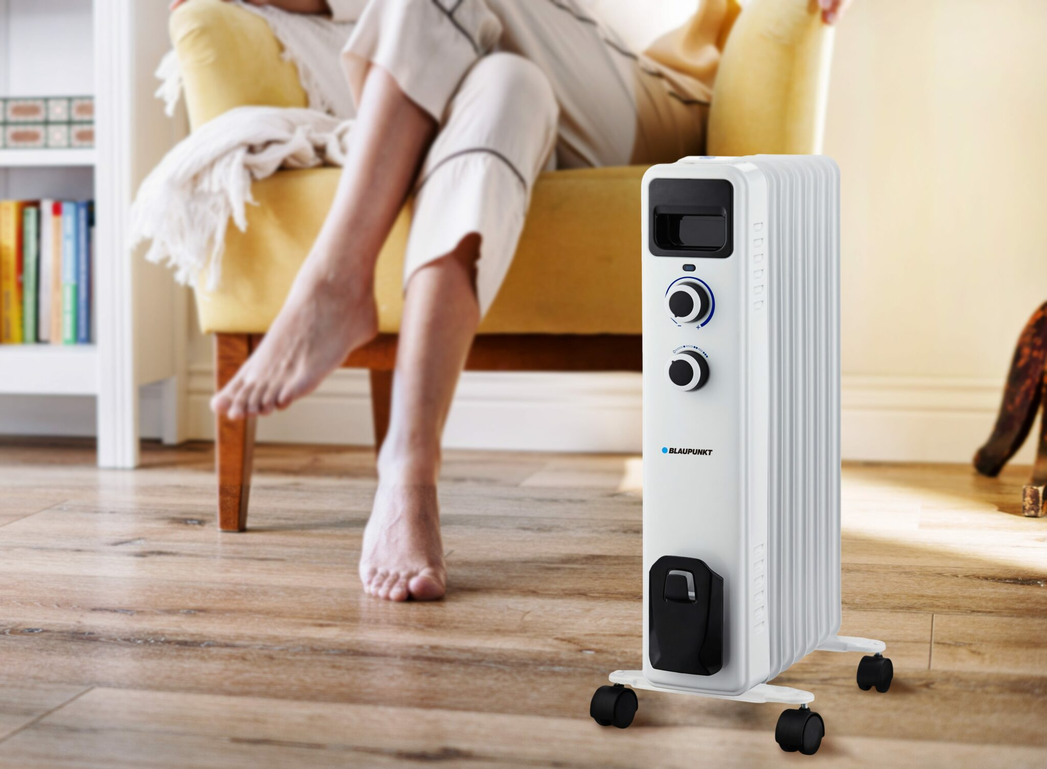 Biały grzejnik olejowy stoi blisko osoby siedzącej na żółtym krześle.