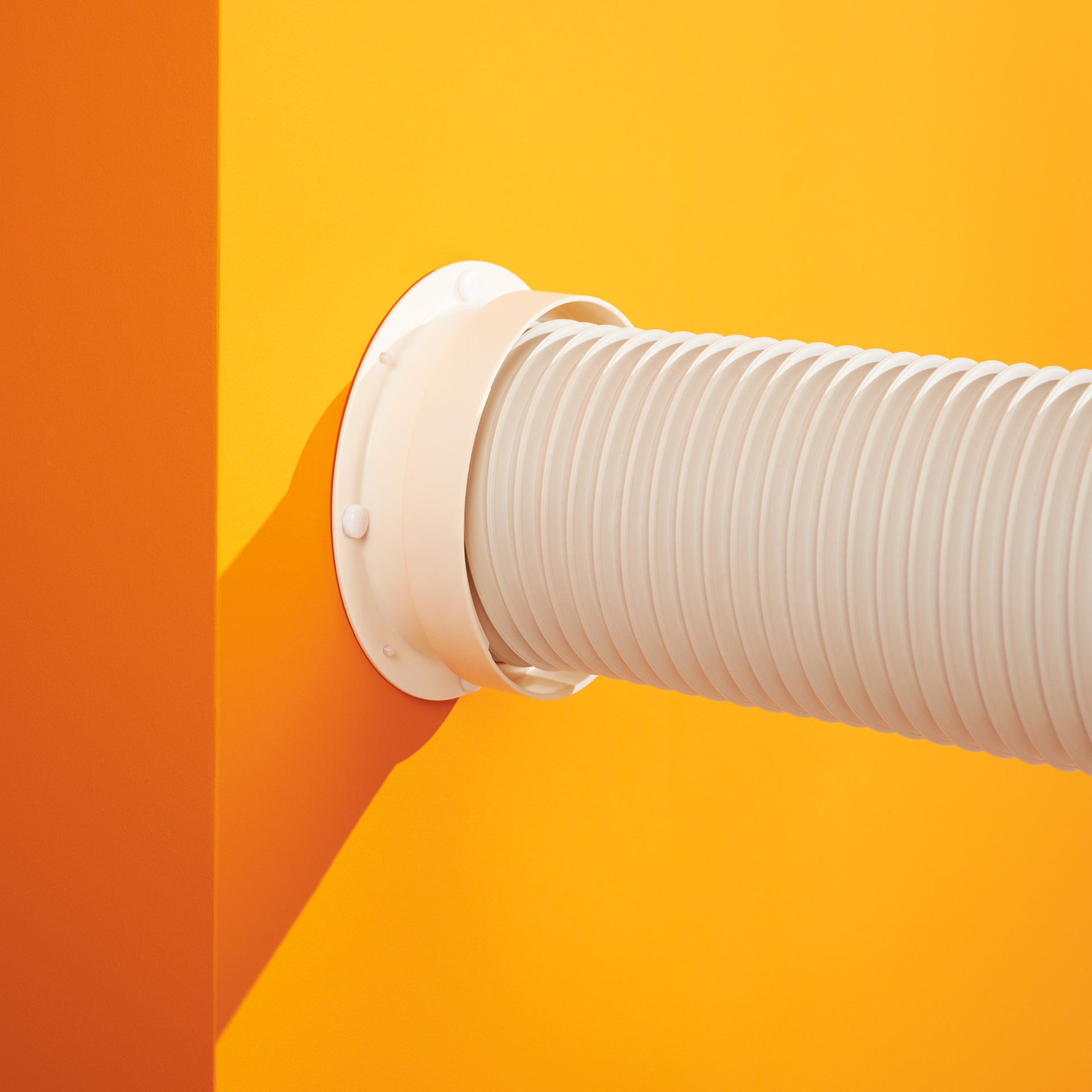 A white plastic ventilation pipe exiting a wall. The wall is painted a bright orange color.