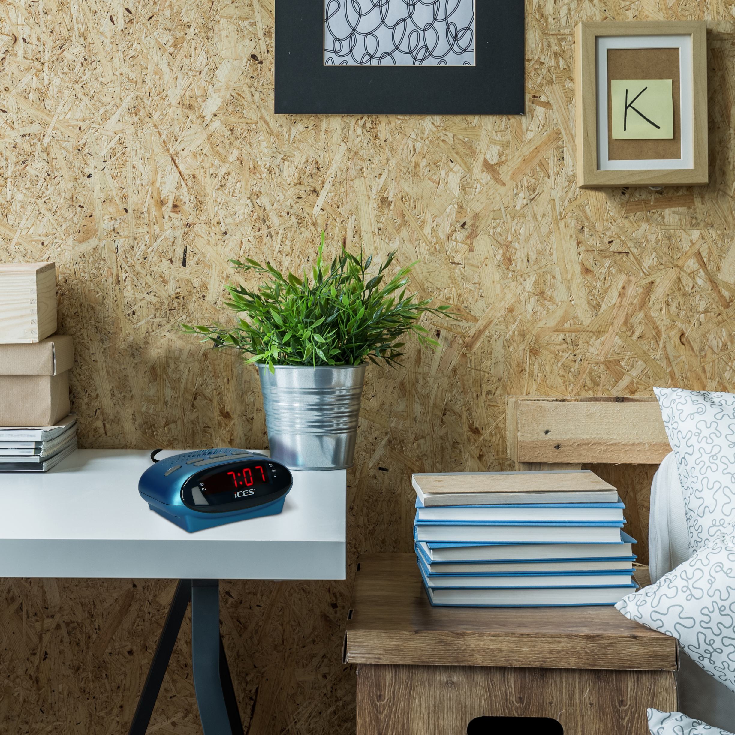 A blue alarm clock on a white desk, with a plant and books. Background has a wooden wall.