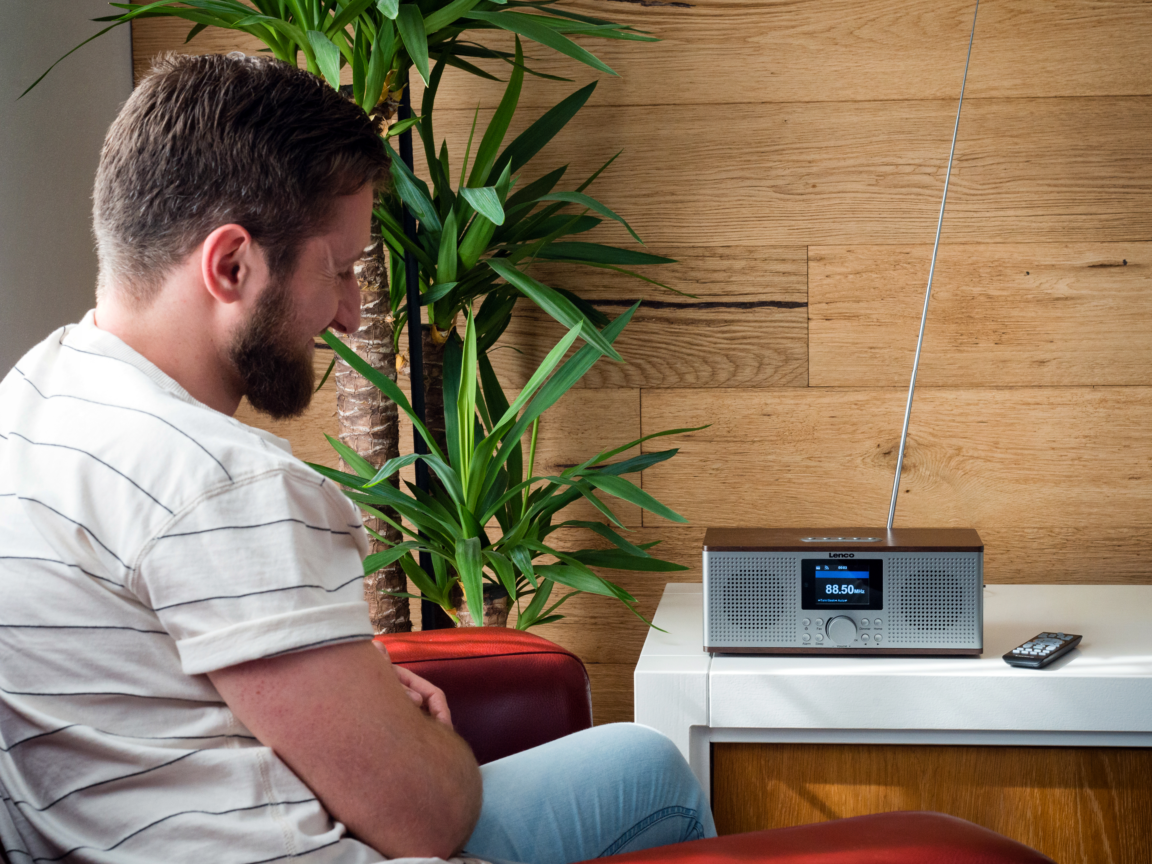 Mężczyzna z brodą siedzi obok radia na białym stole. Roślina i drewniana ściana w tle.