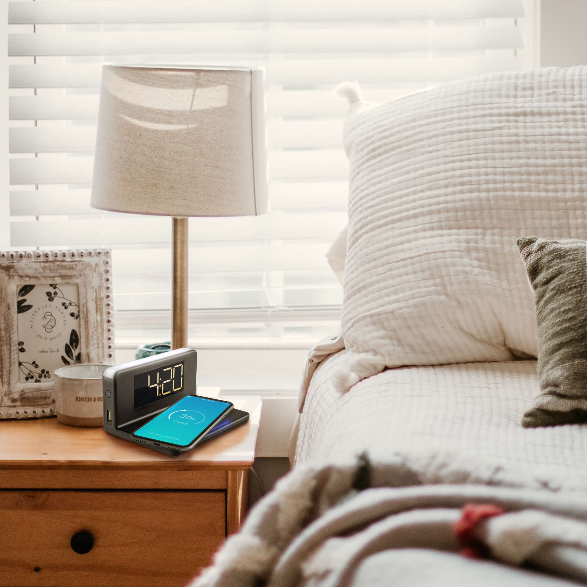 A bedside table with a clock showing 4:20, a phone charging, a lamp, and a white bed.