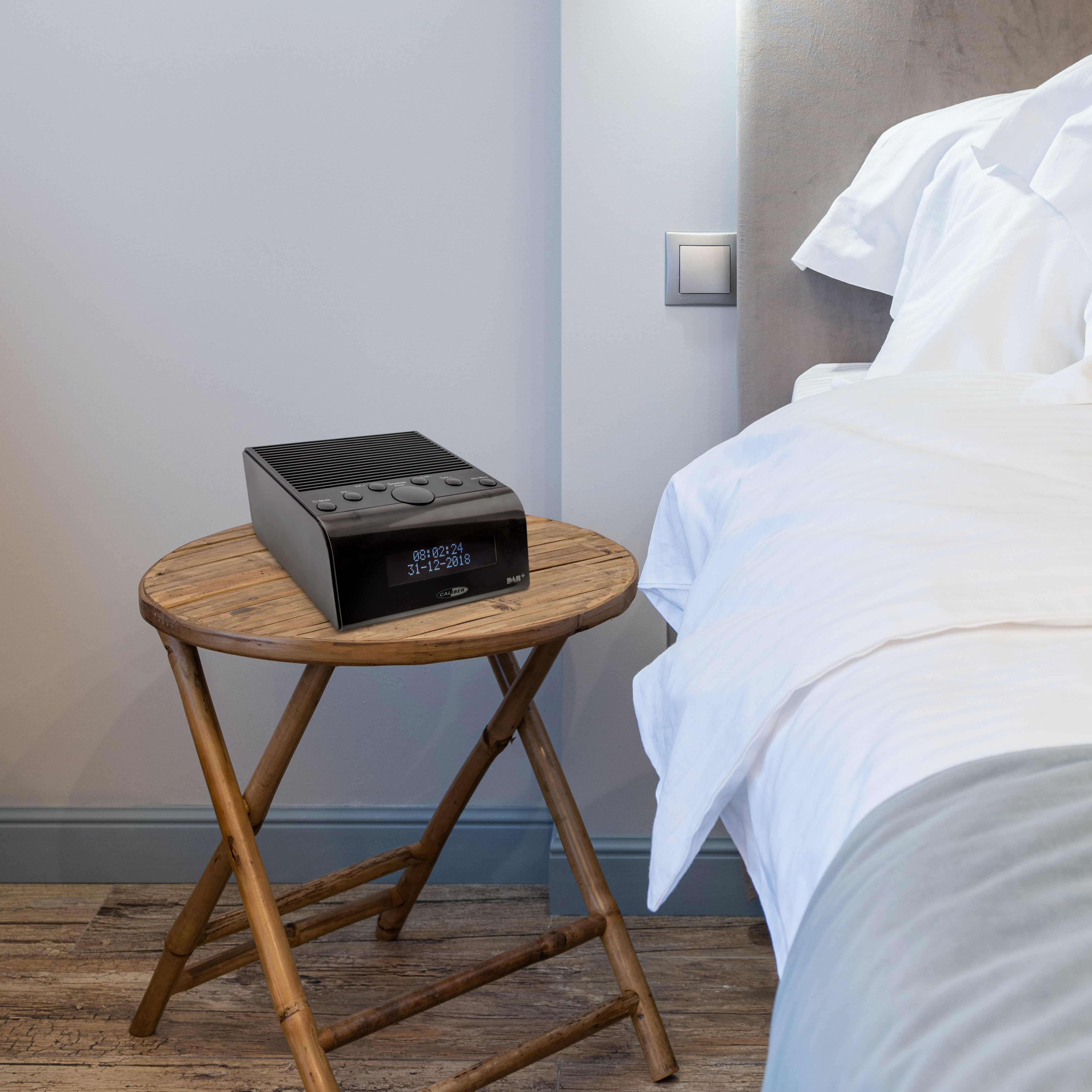 Een zwarte digitale klokradio op een houten tafel naast een bed.