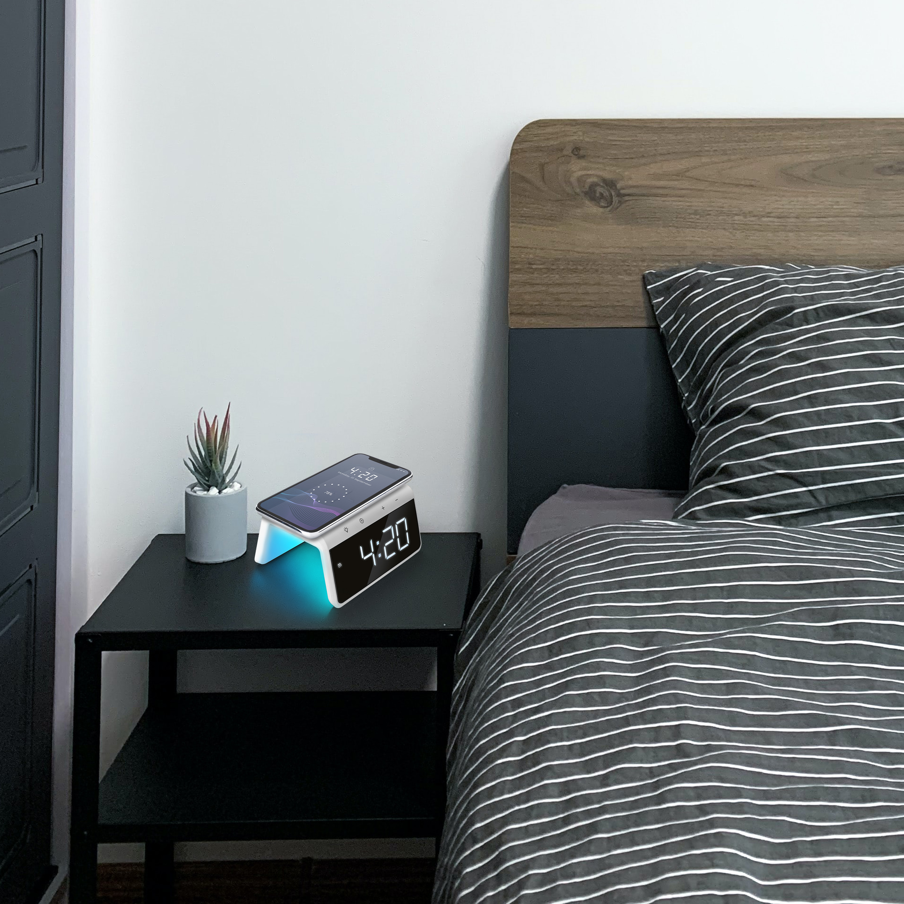 Bedside table with clock/charger and phone. Bed with striped blanket and wooden headboard.
