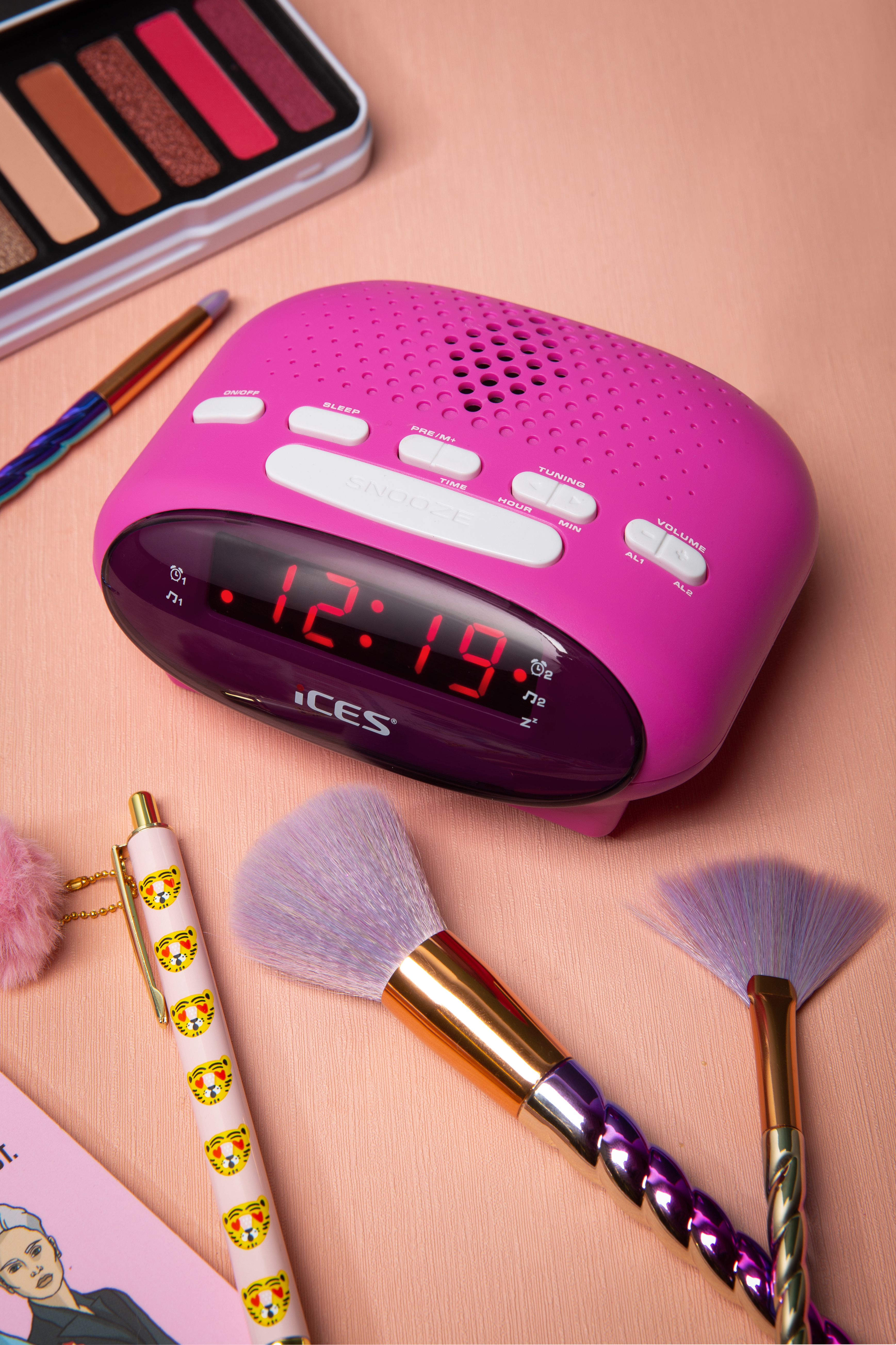 Pink alarm clock with makeup and brushes. Clock shows 12:19. Other items include a pen and eyeshadow.