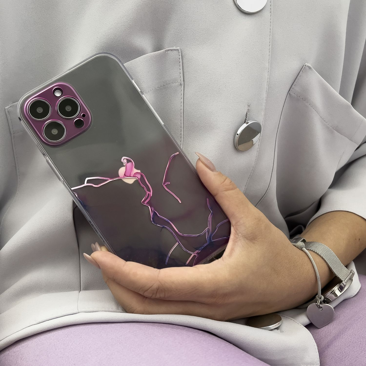 Frau hält Telefon in transparenter Hülle mit rosa Akzenten, trägt graue Jacke und lila Hose.