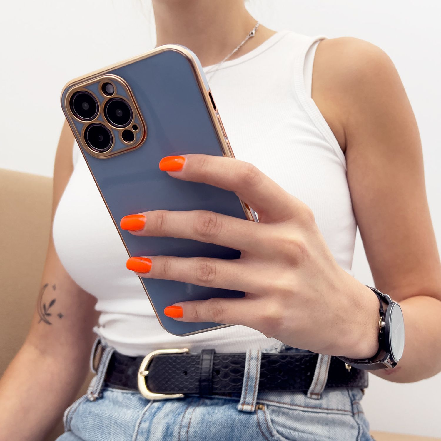Eine Frau hält eine blaue Handyhülle mit orangen Nägeln, ein weißes Oberteil und eine Uhr.