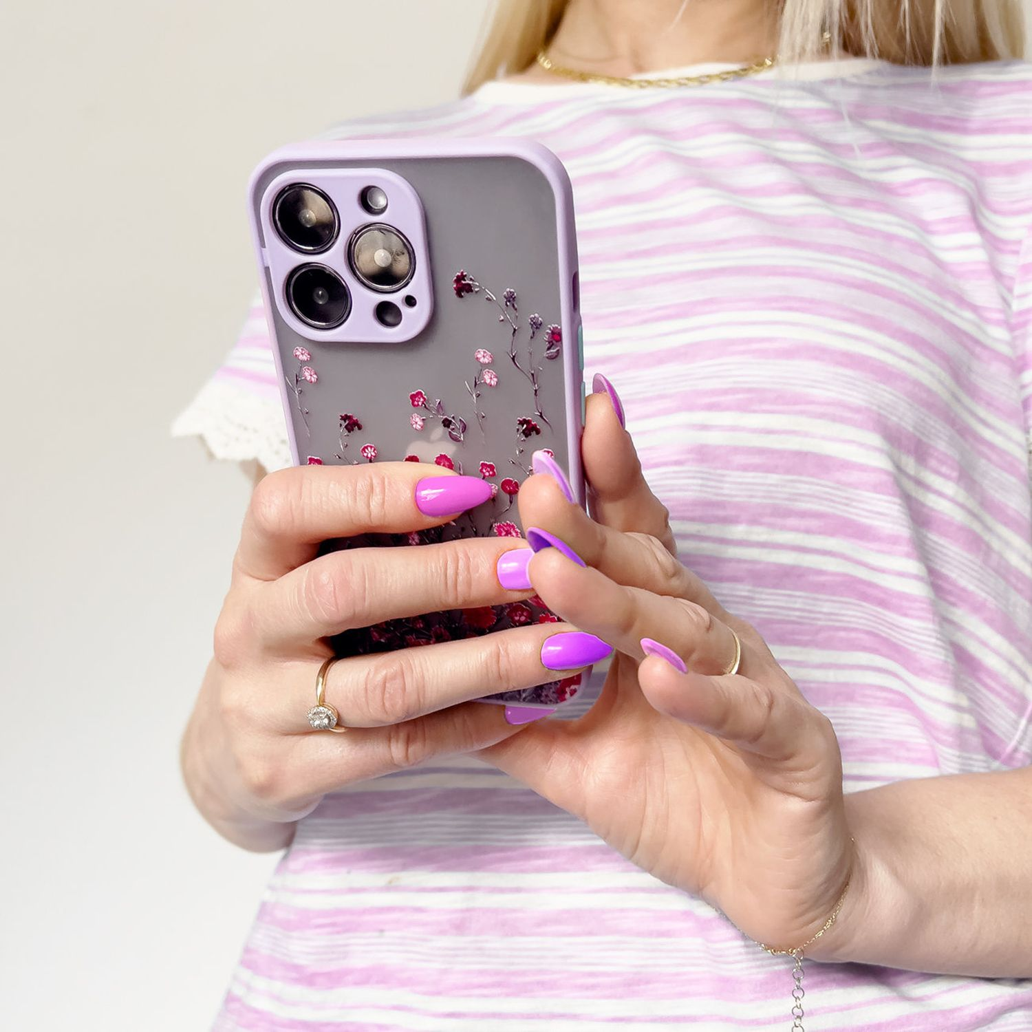 Hände einer Person, die ein Telefon mit lila Hülle und Blumendesign halten. Die Nägel sind lila lackiert.