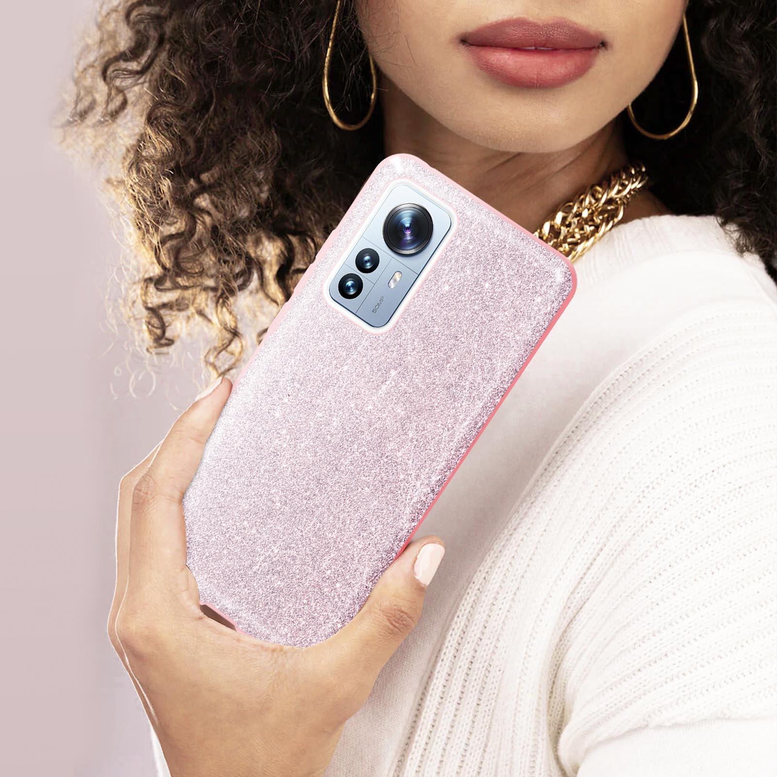 Donna tiene telefono rosa glitterato, collana dorata e maglione bianco.