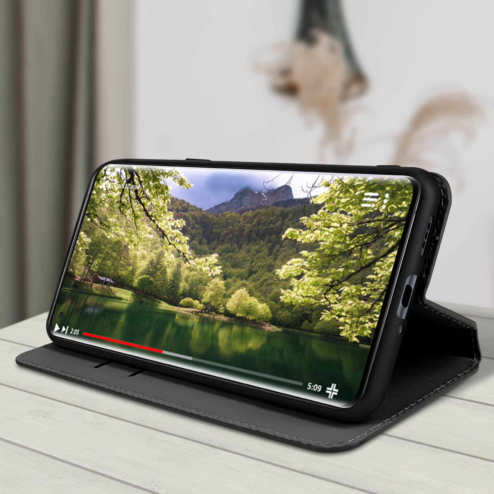 Een telefoon in een zwarte hoes speelt een video van een meer af. De hoes ligt op een houten tafel.