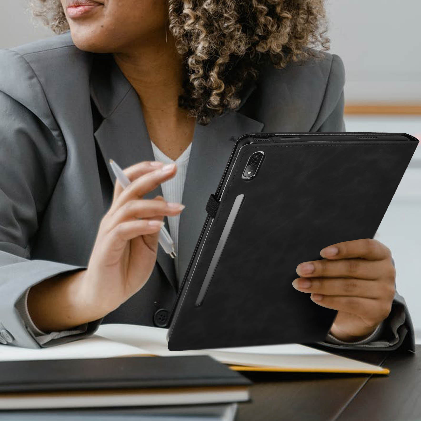 Donna in completo grigio che tiene un tablet nero con pennino. Libri visibili sullo sfondo.