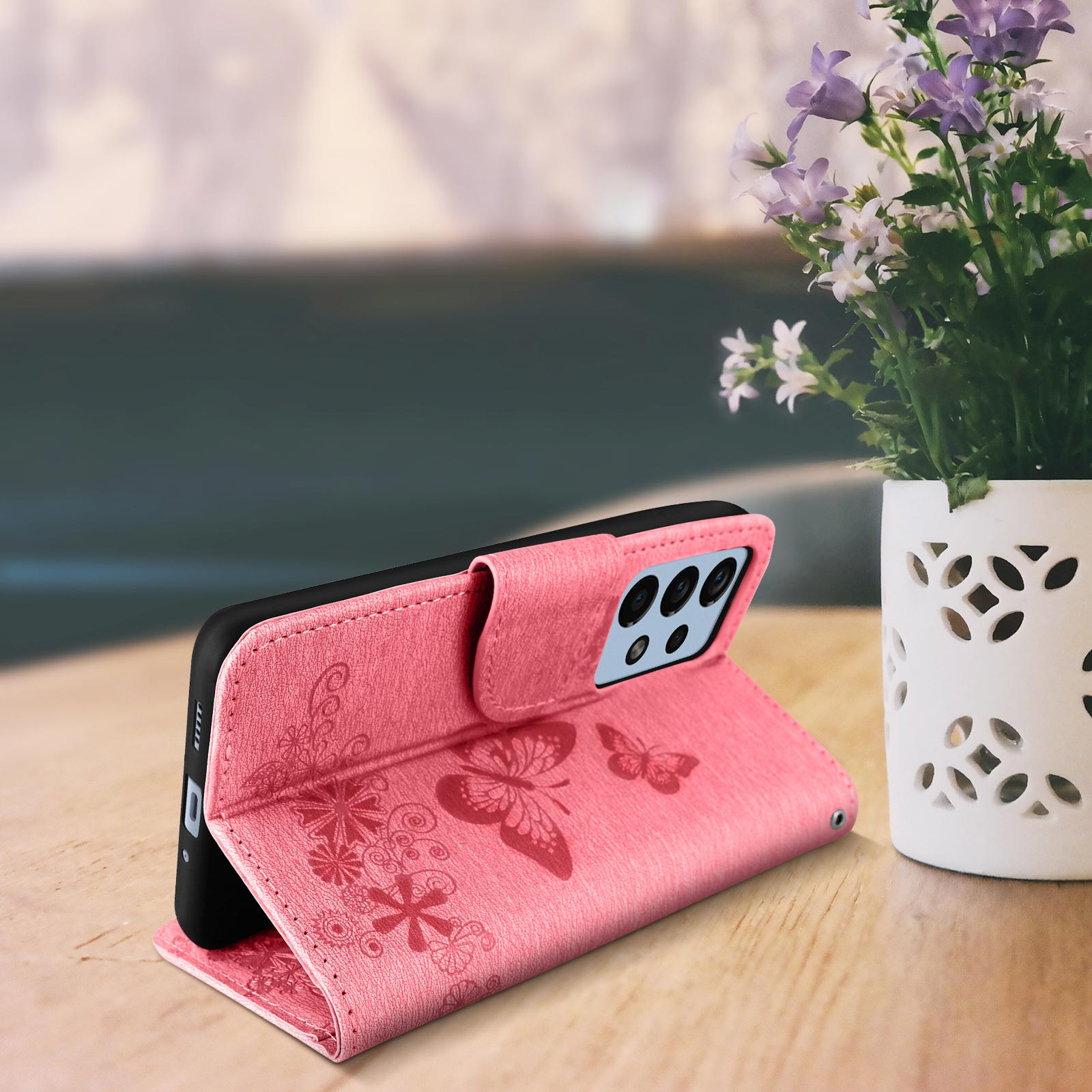 Una custodia rosa aperta con telefono dentro e fiori sullo sfondo, su un tavolo di legno.