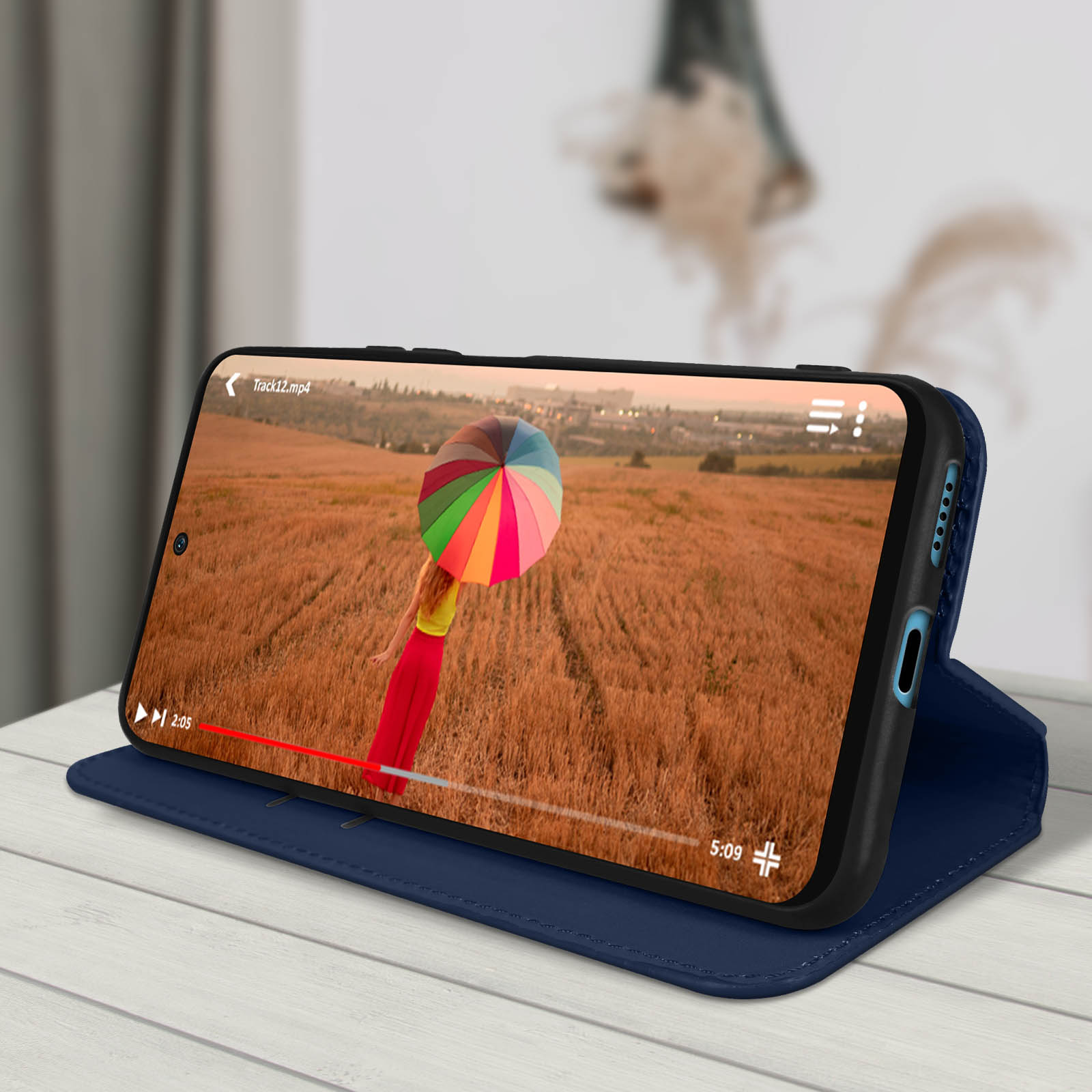 Téléphone affichant vidéo d'une personne avec parapluie dans champ. Téléphone dans étui bleu sur table.