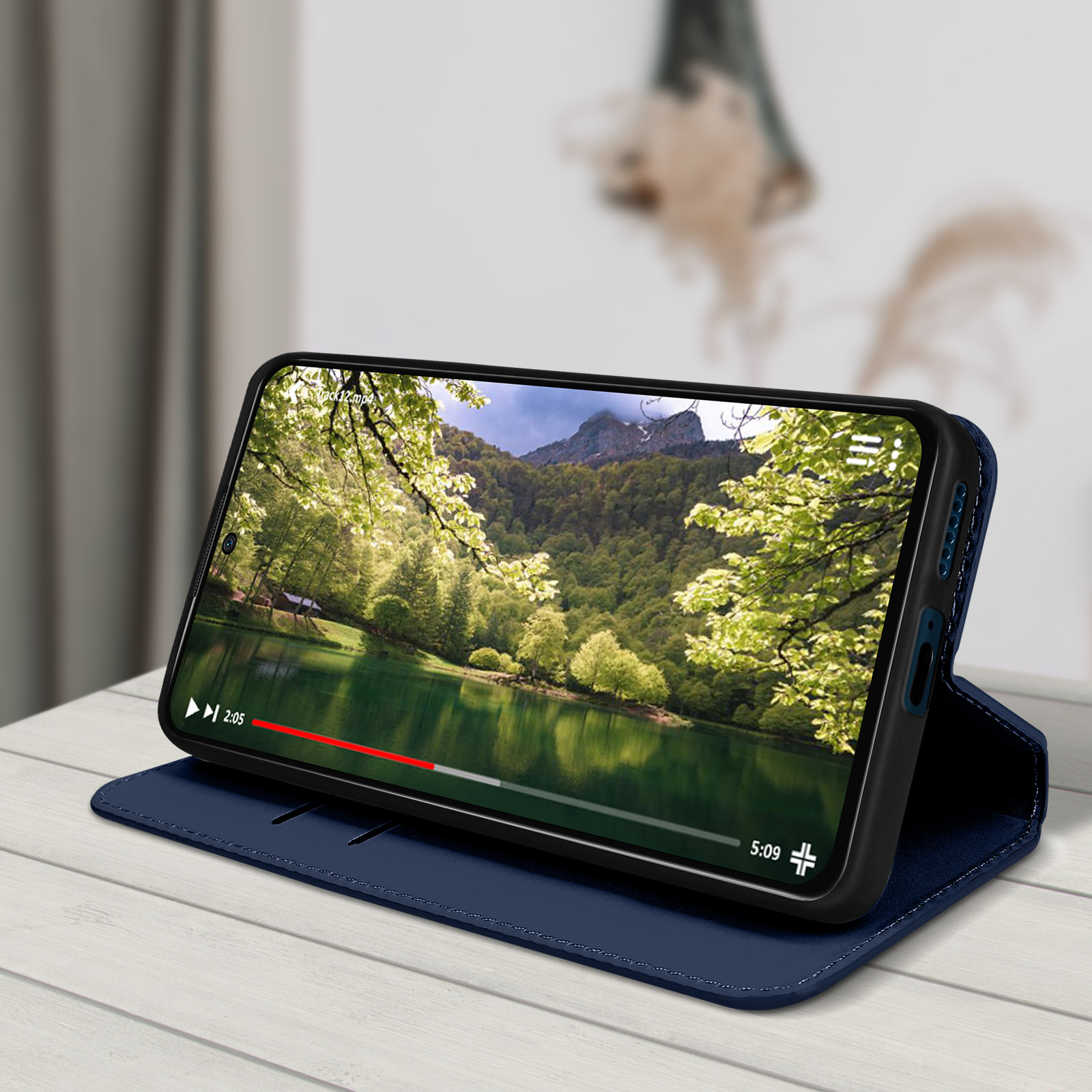 Telefono su custodia blu scuro, riproduzione vista lago, tavolo in legno bianco.